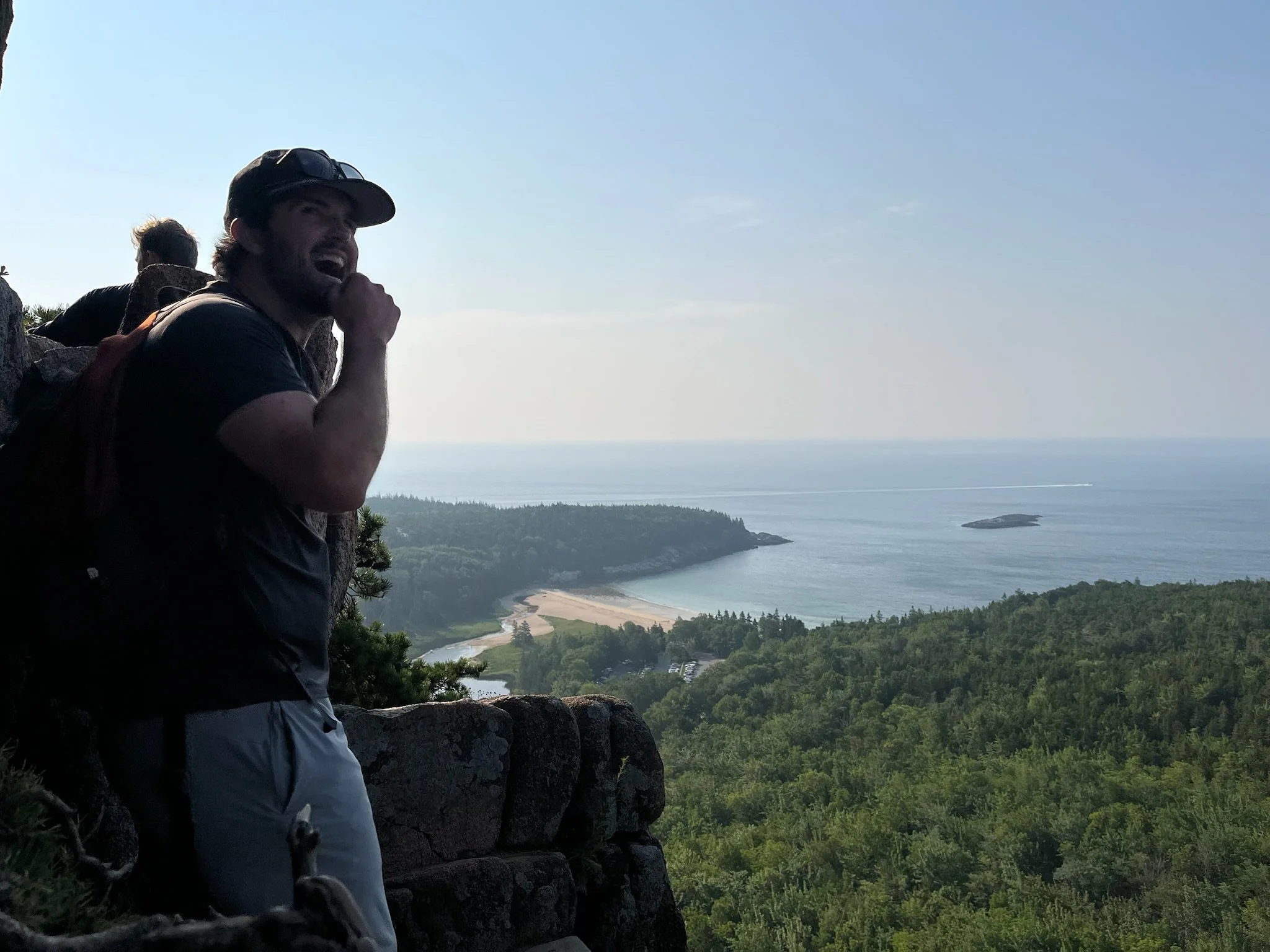 A man with a backpack on a mountain overlook, smiling and resting his chin on his hand, with a scenic view of ocean, islands, and greenery in the background.