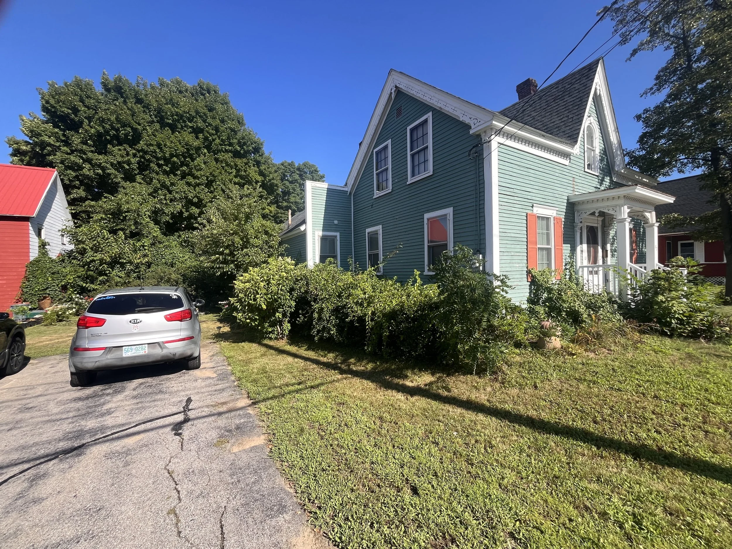 A light blue house with white trim and a front porch, surrounded by green shrubs and trees under a clear blue sky. A car is parked in the driveway.