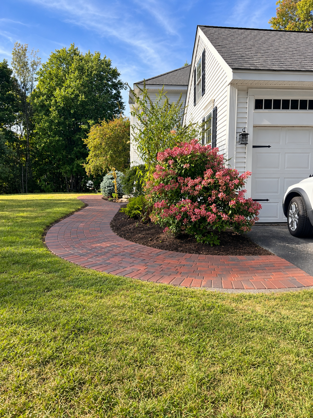 Curved brick walkway in front of a white house with black shutters, pink flowering bush, green trees, and a parked white car on a sunny day.