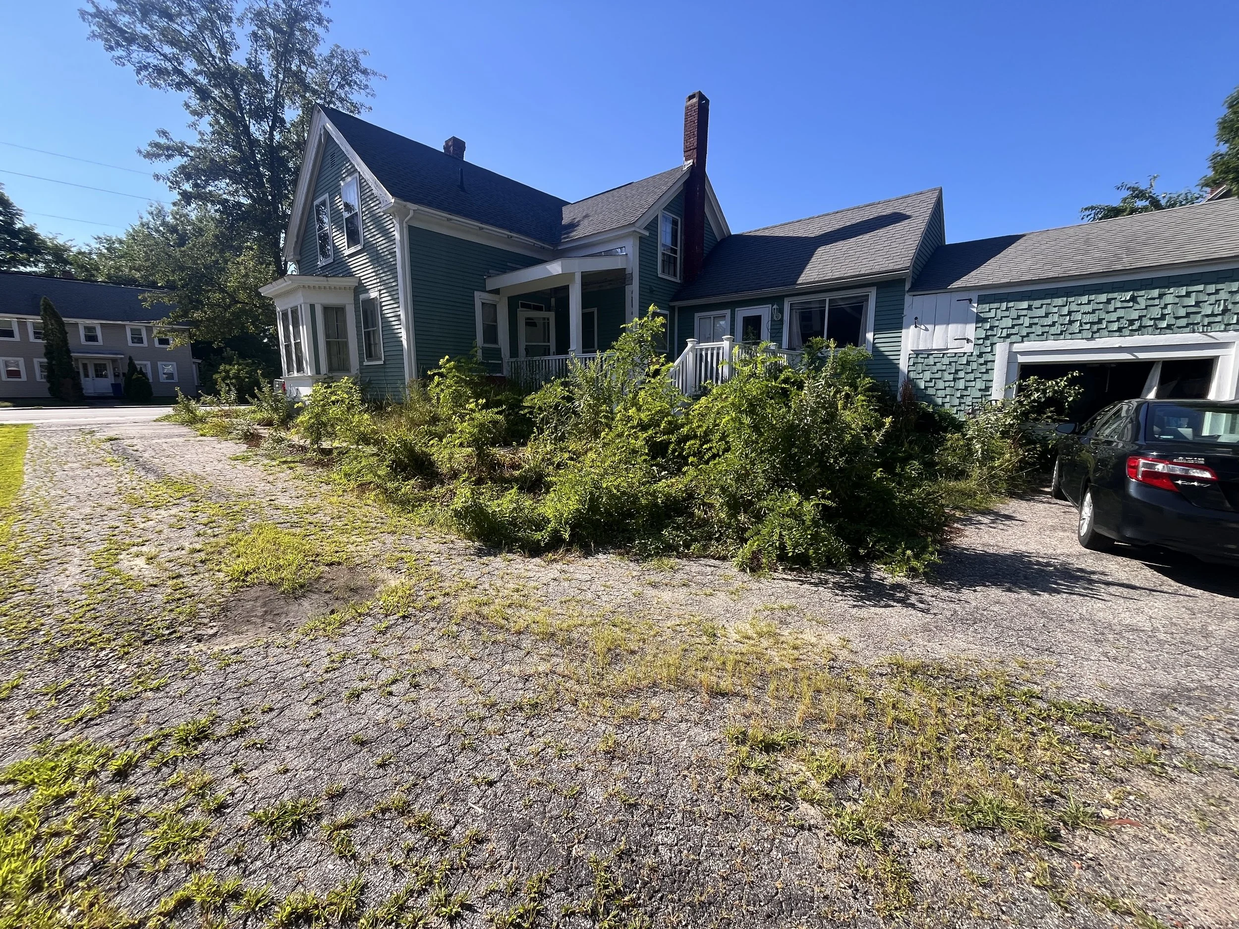 A house with teal siding, a brick chimney, and a white porch with steps. There is a car parked in the driveway and overgrown grass and plants around the house under a clear blue sky.