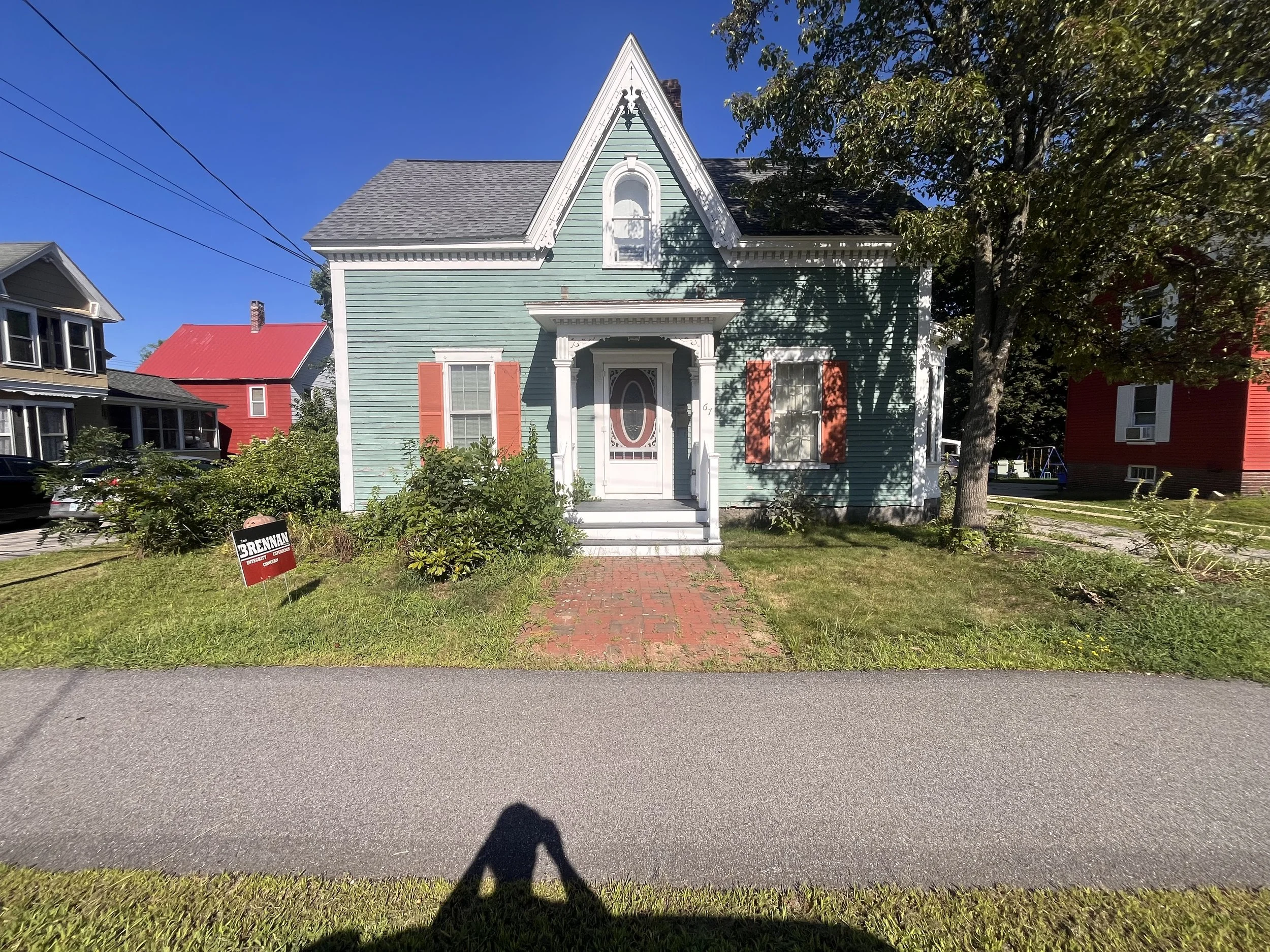 Colorful house with a teal exterior and red shutters, front steps leading to a decorative front door, surrounding greenery, and a large tree on the right side under a clear blue sky.