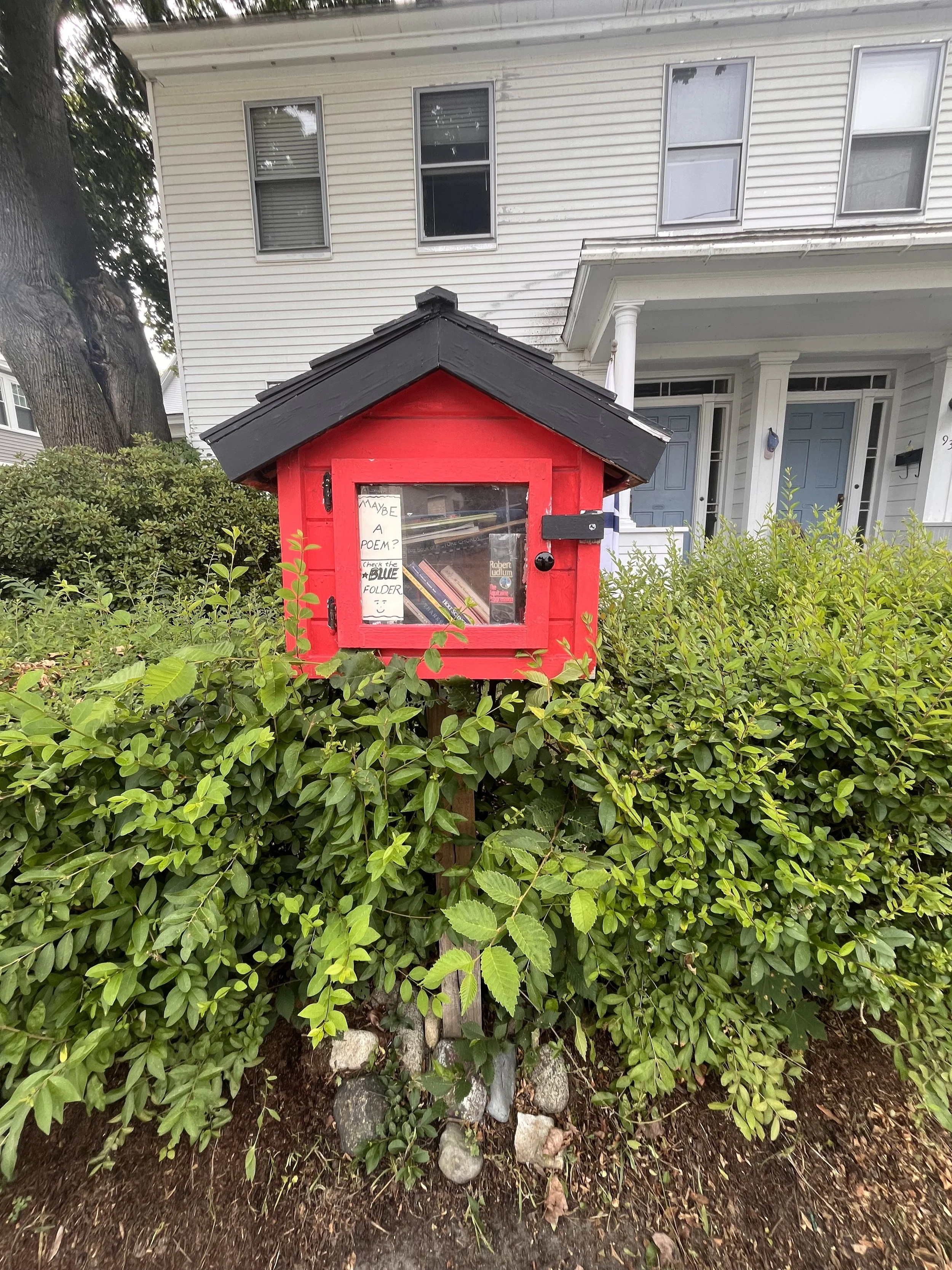 A red mailbox with a small window displaying books and notes, situated in a garden in front of a white house with blue doors and several windows.