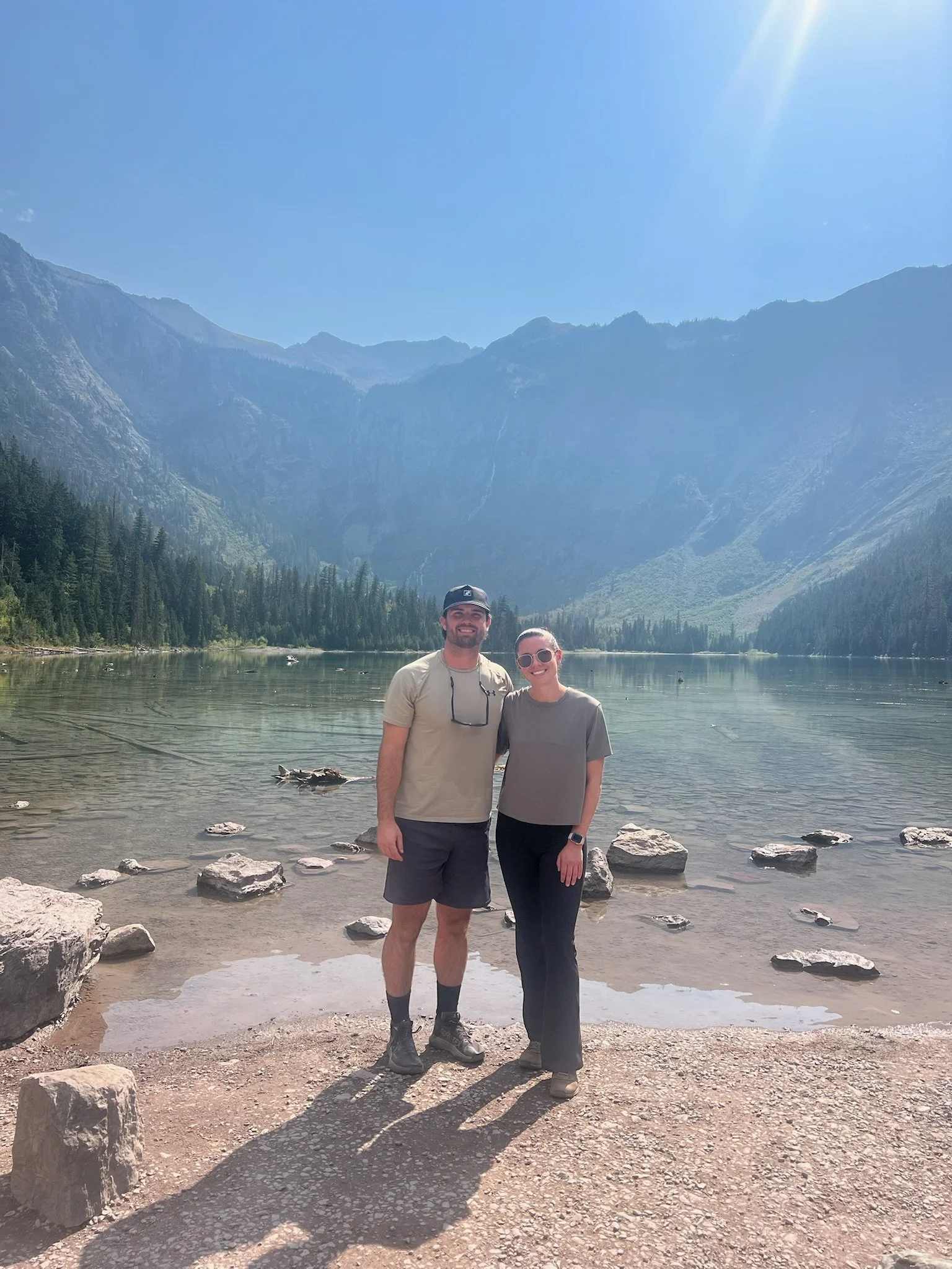 A couple standing on a rocky shore in front of a lake with mountains in the background, smiling, under a bright sunny sky.