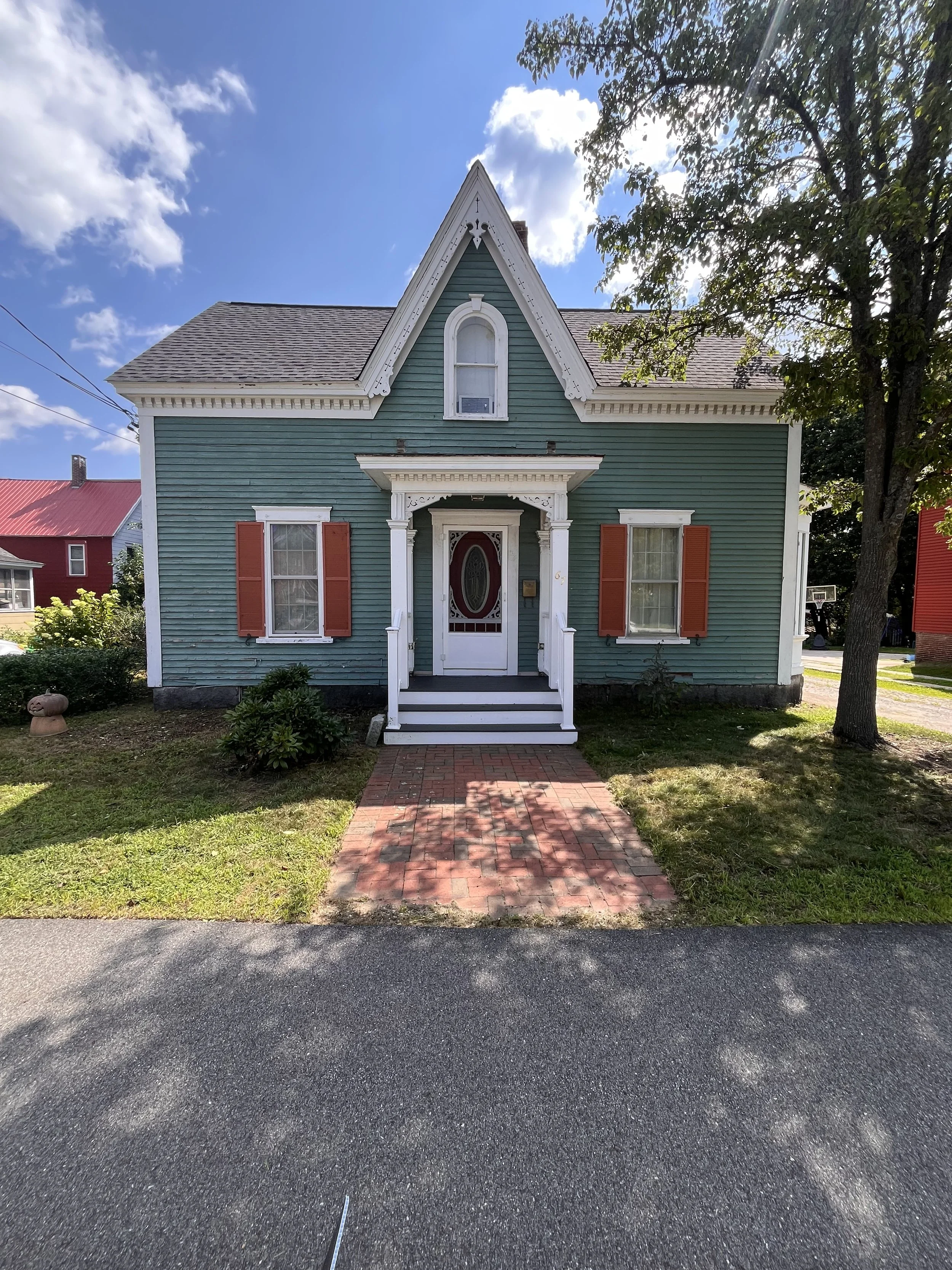 A two-story house painted in teal with white trim, red shutters, a covered porch, and a small window with an air conditioning unit. The house is surrounded by trees and a lawn, with a brick walkway leading to the front door.