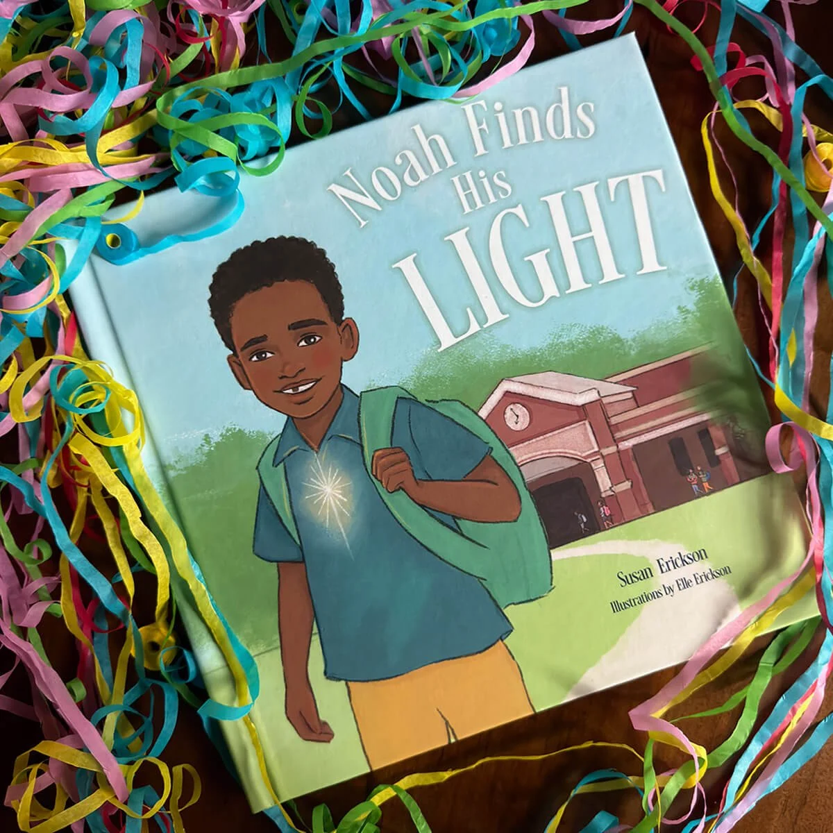 A children's book titled "Noah Finds His Light" by Susan Erickson, with illustrations by Elle Erickson, is surrounded by colorful paper confetti.