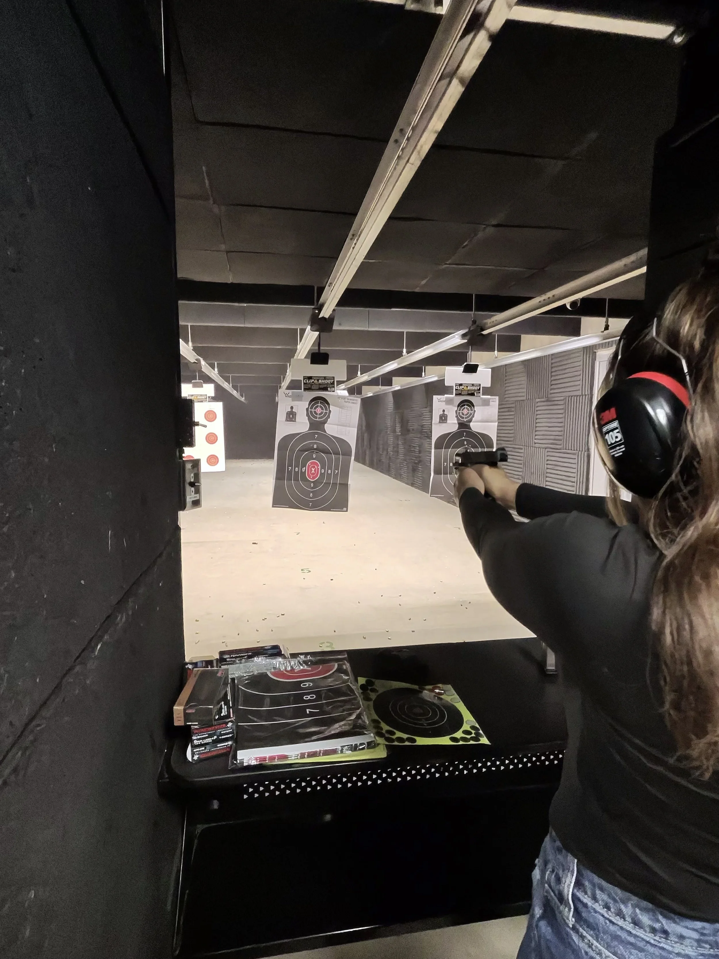 An indoor shooting range with targets hanging from the ceiling, a person wearing ear protection aiming a firearm, and shelves with ammunition and target sheets.