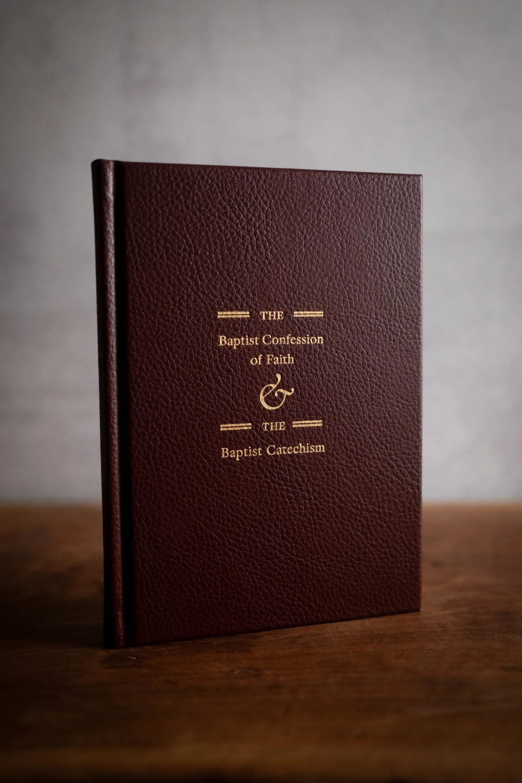 A maroon-colored textured hardcover book titled 'The Baptist Confession of Faith & The Baptist Catechism' standing upright on a wooden surface against a plain background.