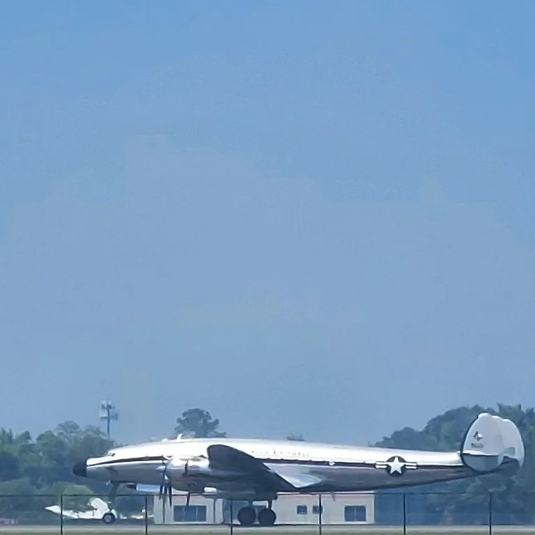 A vintage aircraft parked on an airfield with a clear blue sky and some distant trees in the background.