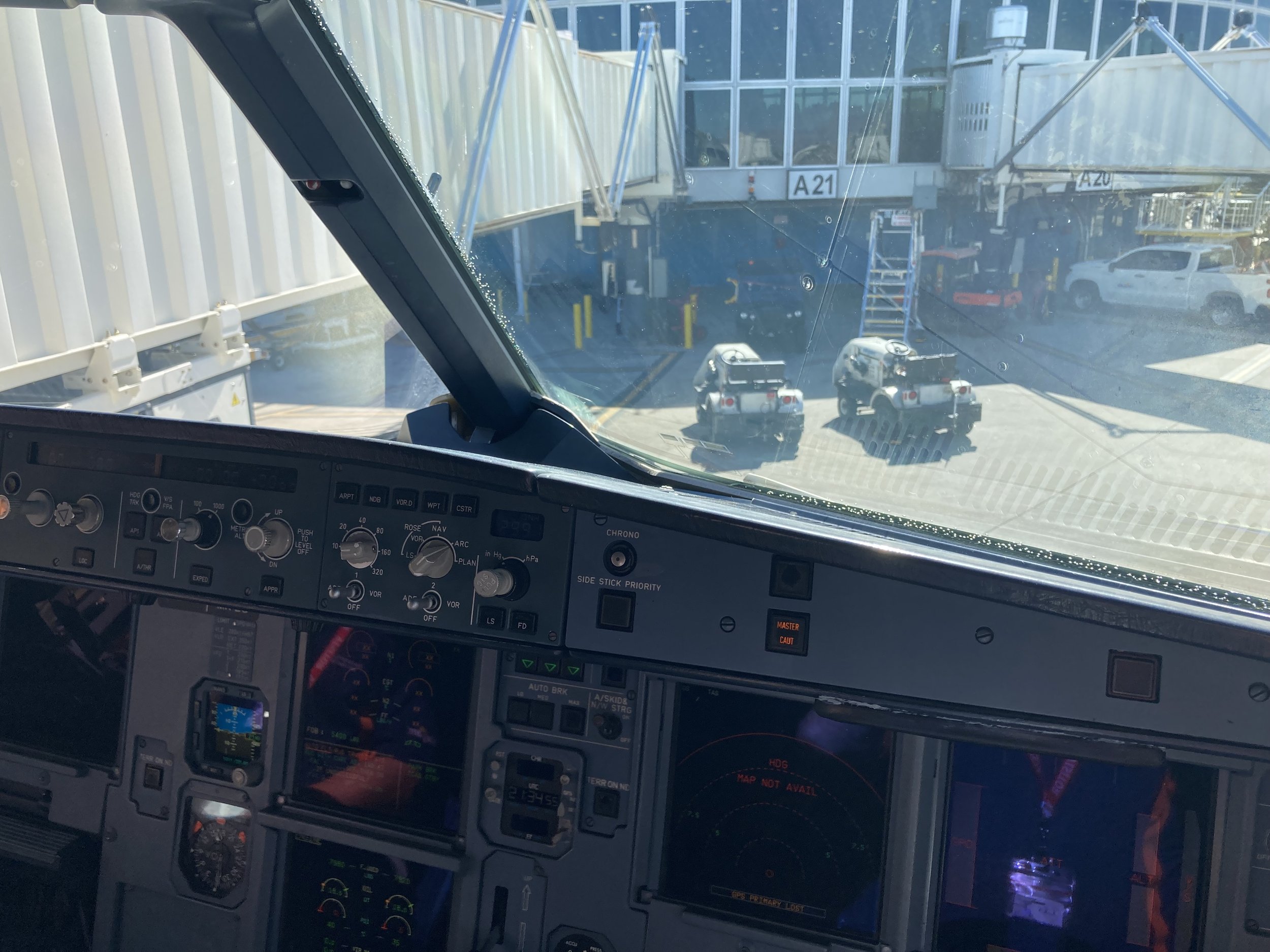 Photo from the cockpit of an airplane showing various instrument panels and controls. Outside the cockpit window, a jet bridge connected to the aircraft with a white car and airport ground equipment visible in the background.