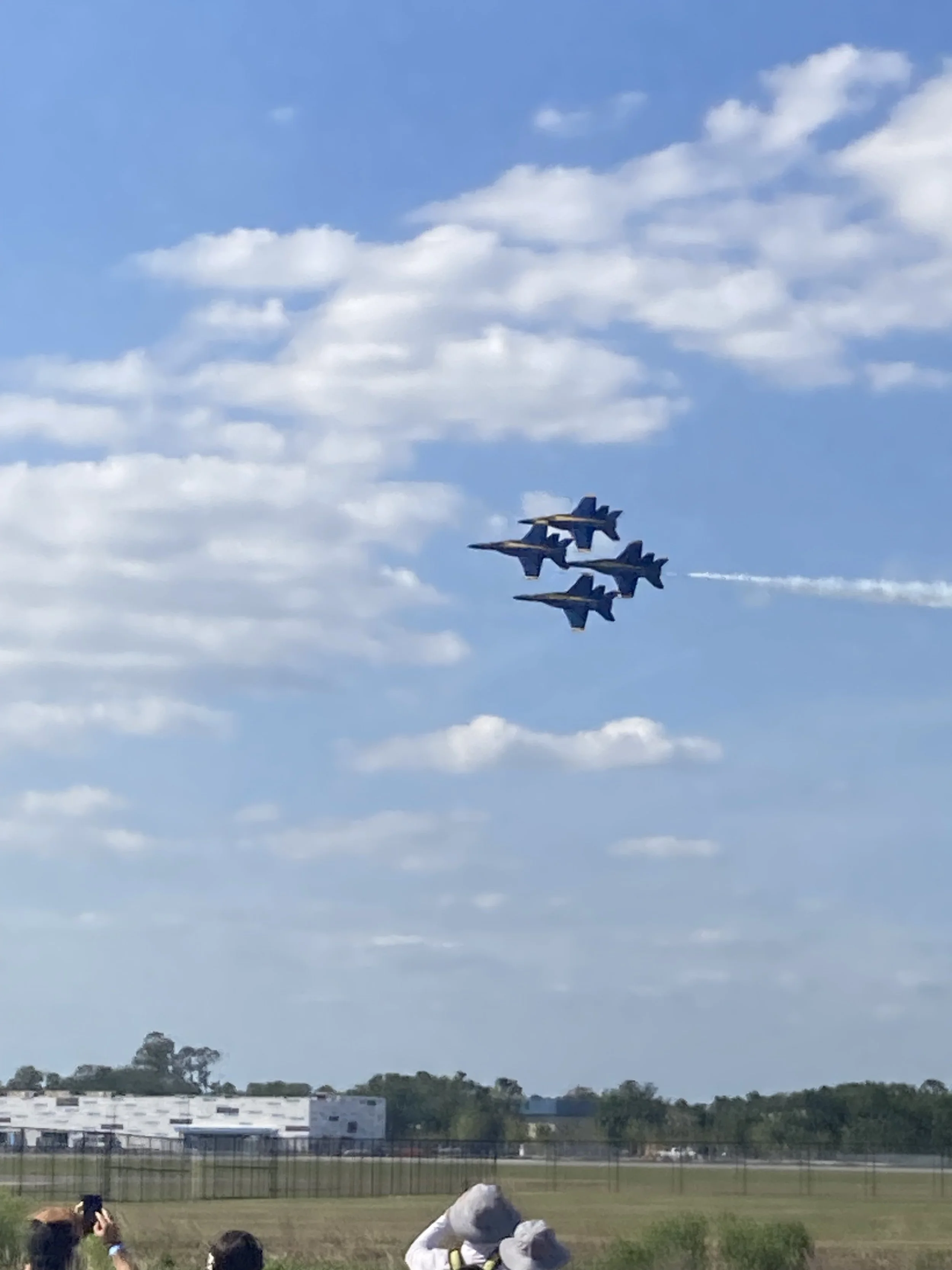 Four fighter jets flying in a tight formation through a partly cloudy sky, with spectators taking photos in the foreground and a landscape with trees and a white building below.