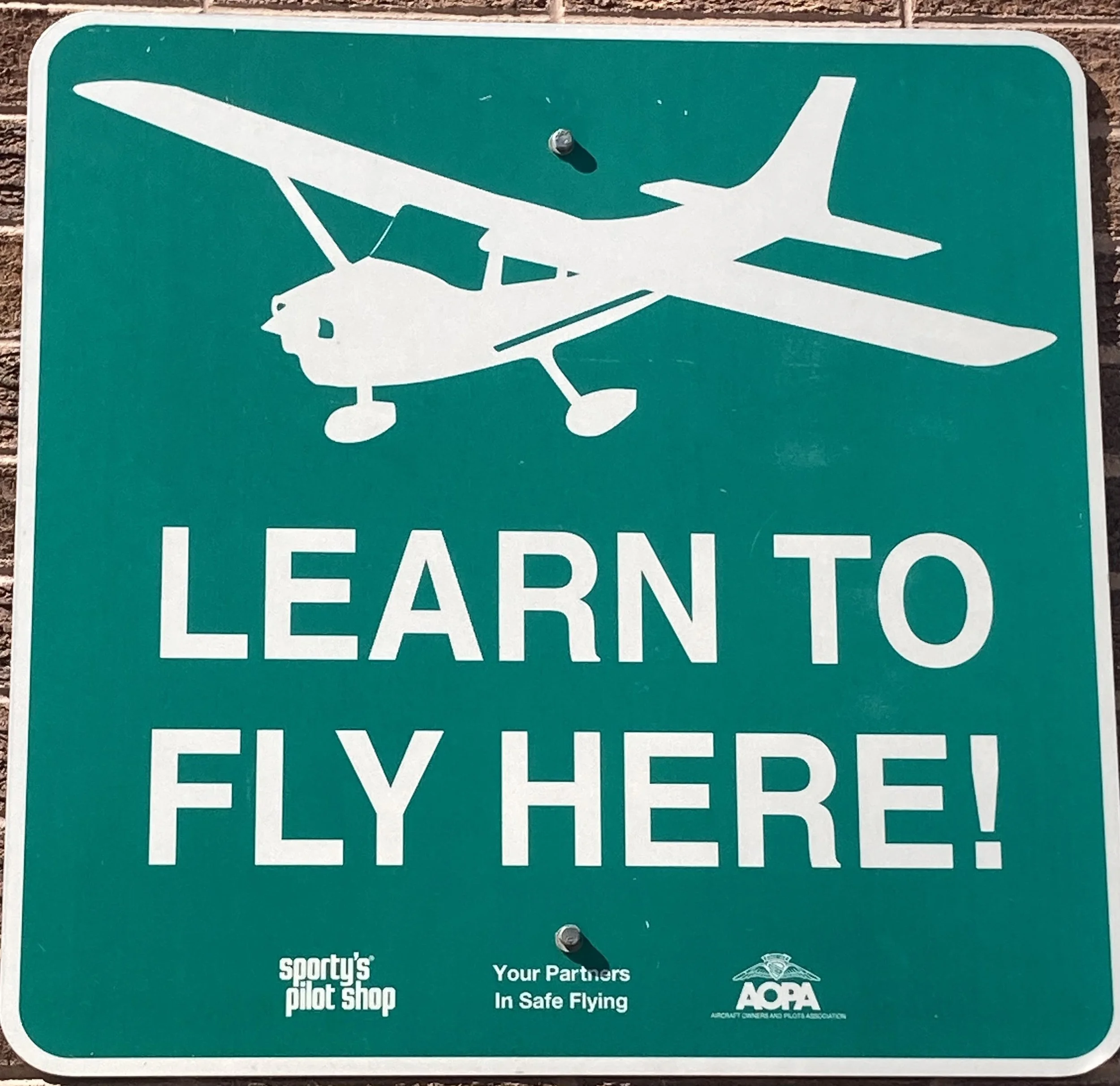 Green sign with a white airplane and the words "LEARN TO FLY" signified as a pilot training area, with additional small text at the bottom mentioning the sport's pilot school, safe flying, and the Aero Club.