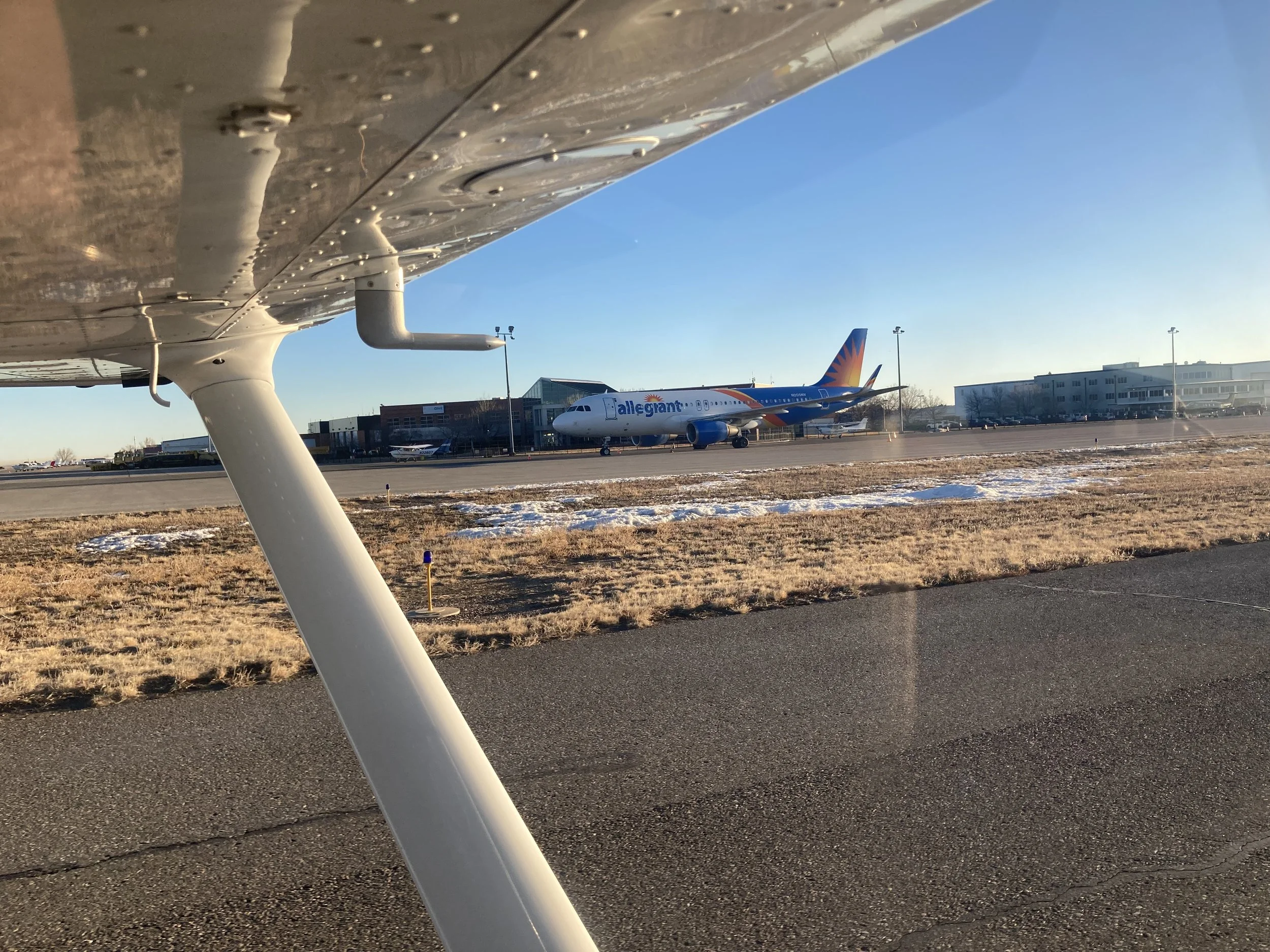 View of an airport runway with a parked Allegiant airplane in the background, captured from the wing of a small aircraft, under a clear blue sky with some buildings and other aircraft nearby.