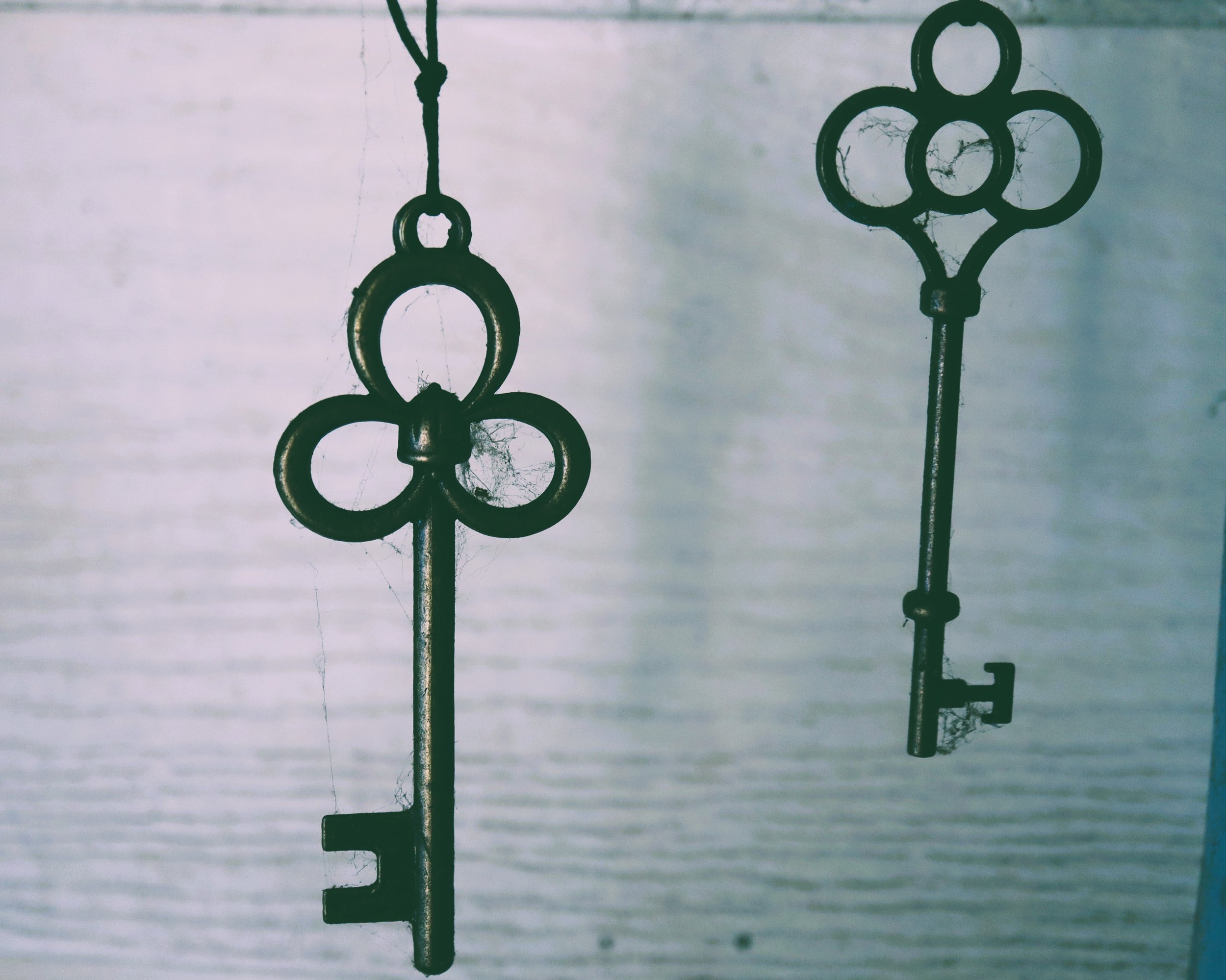 Two old, rusty keys hanging from a surface with cobwebs, against a cloudy, overcast sky.