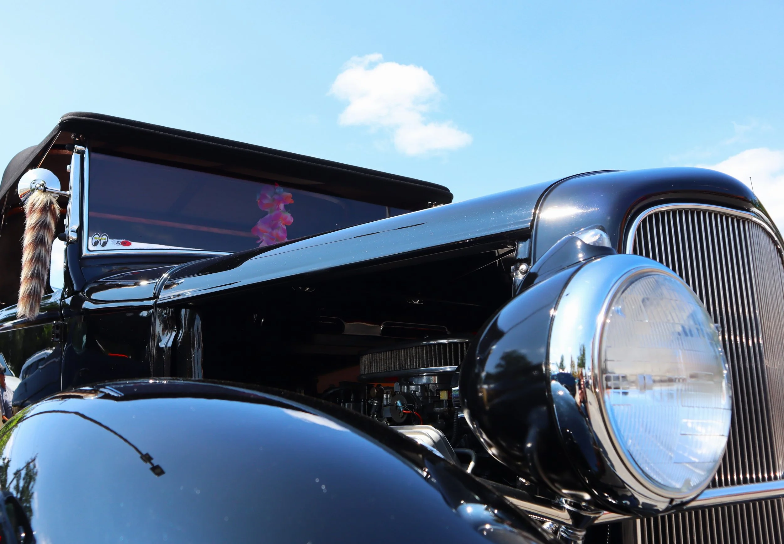 Close-up of a vintage black car with chrome details, partially visible under a bright blue sky with white clouds.