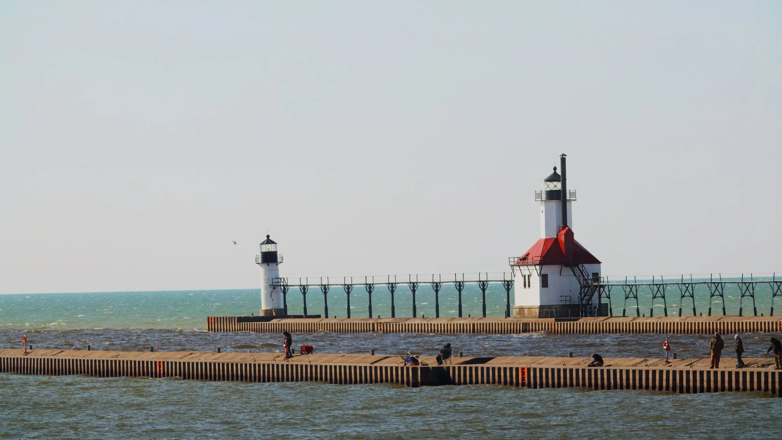 A pier with two lighthouses extending into the water, with people walking and fishing on the pier under a cloudy sky.