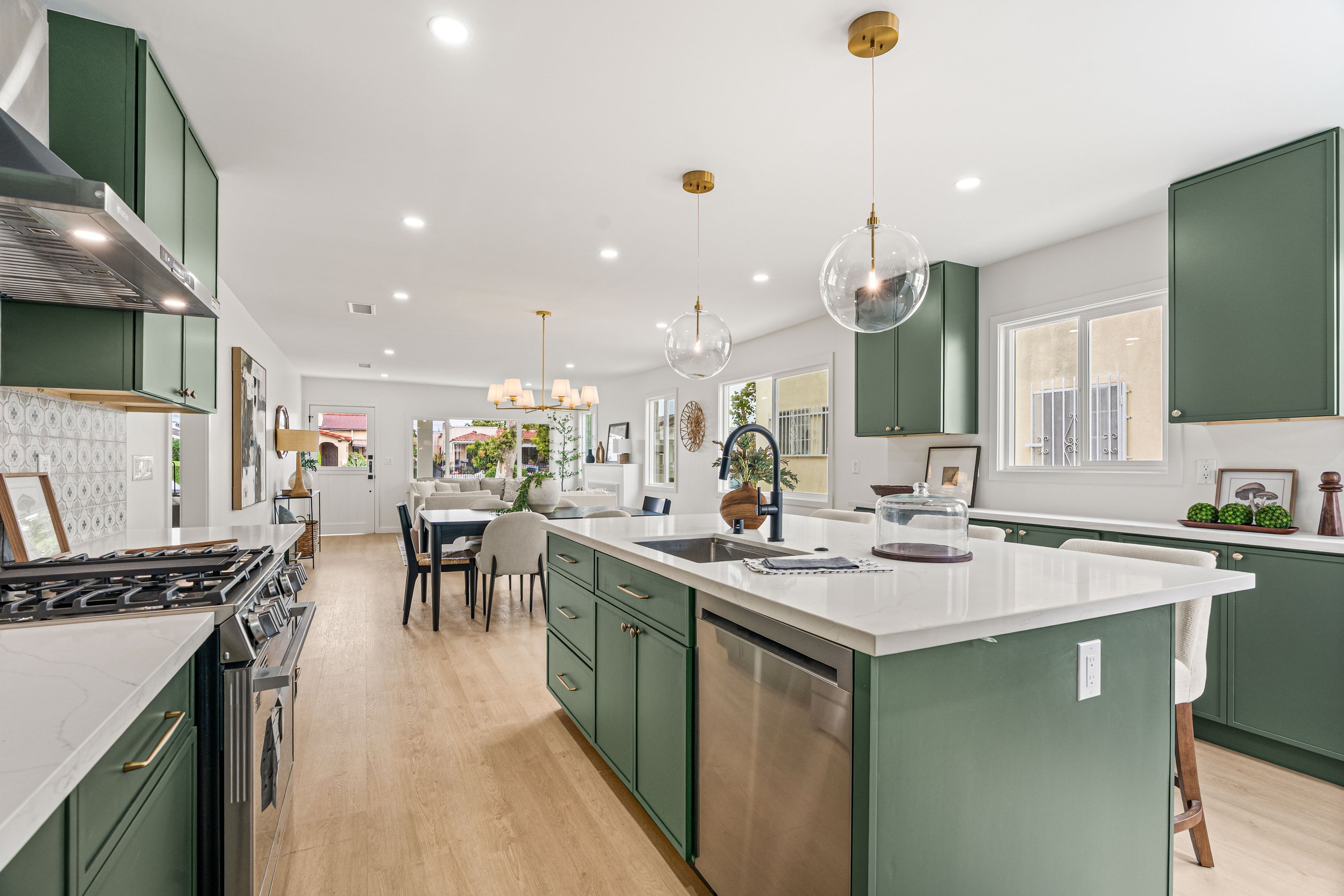 Modern kitchen with green cabinets, white countertops, stainless steel appliances, pendant lights, and open layout with dining and living areas visible.