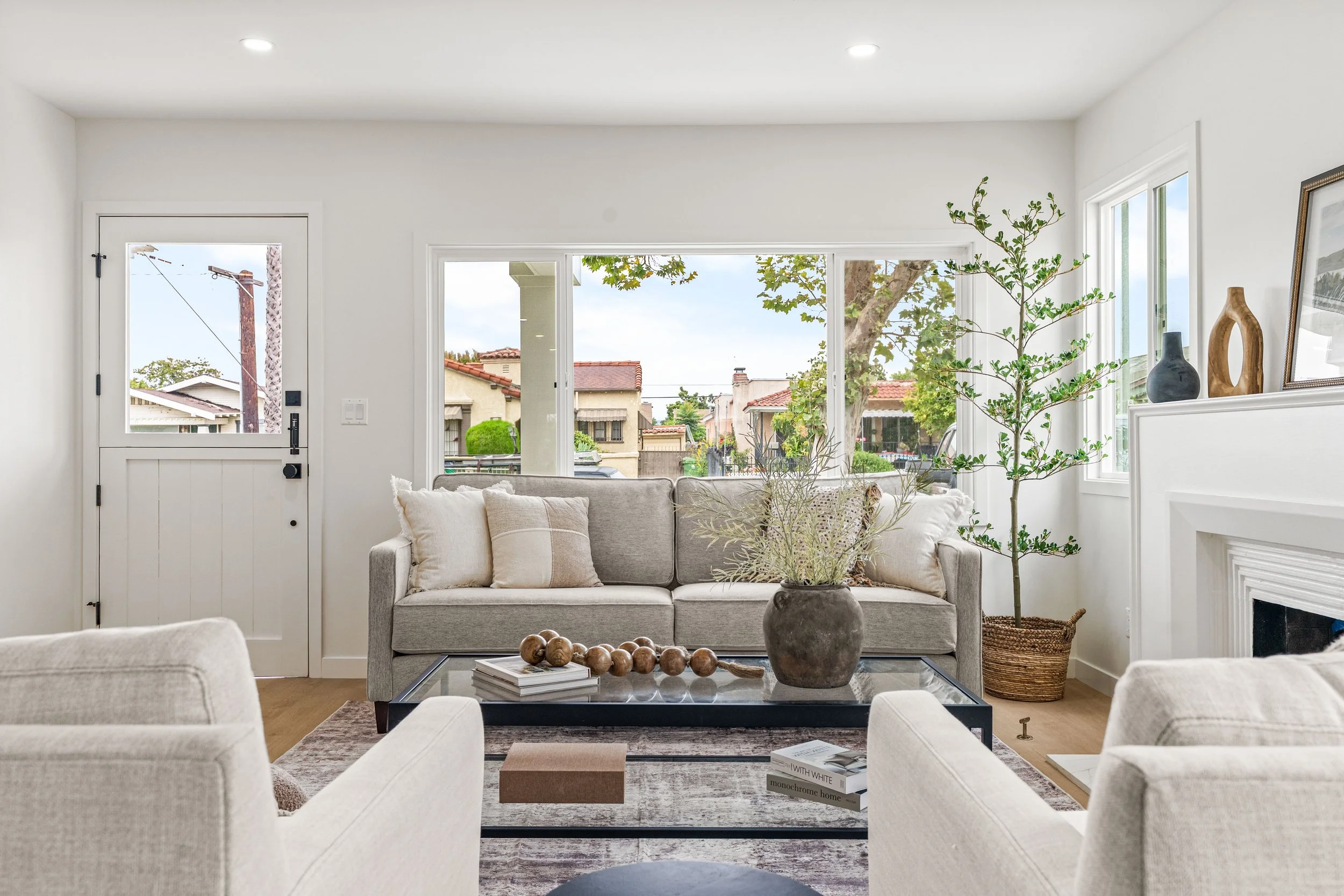 A modern, bright living room with white walls and large windows showing trees and houses outside. There is a light gray sofa with pillows, a black coffee table with books and decorative objects, and a potted plant in a dark pot. Decor items like vase