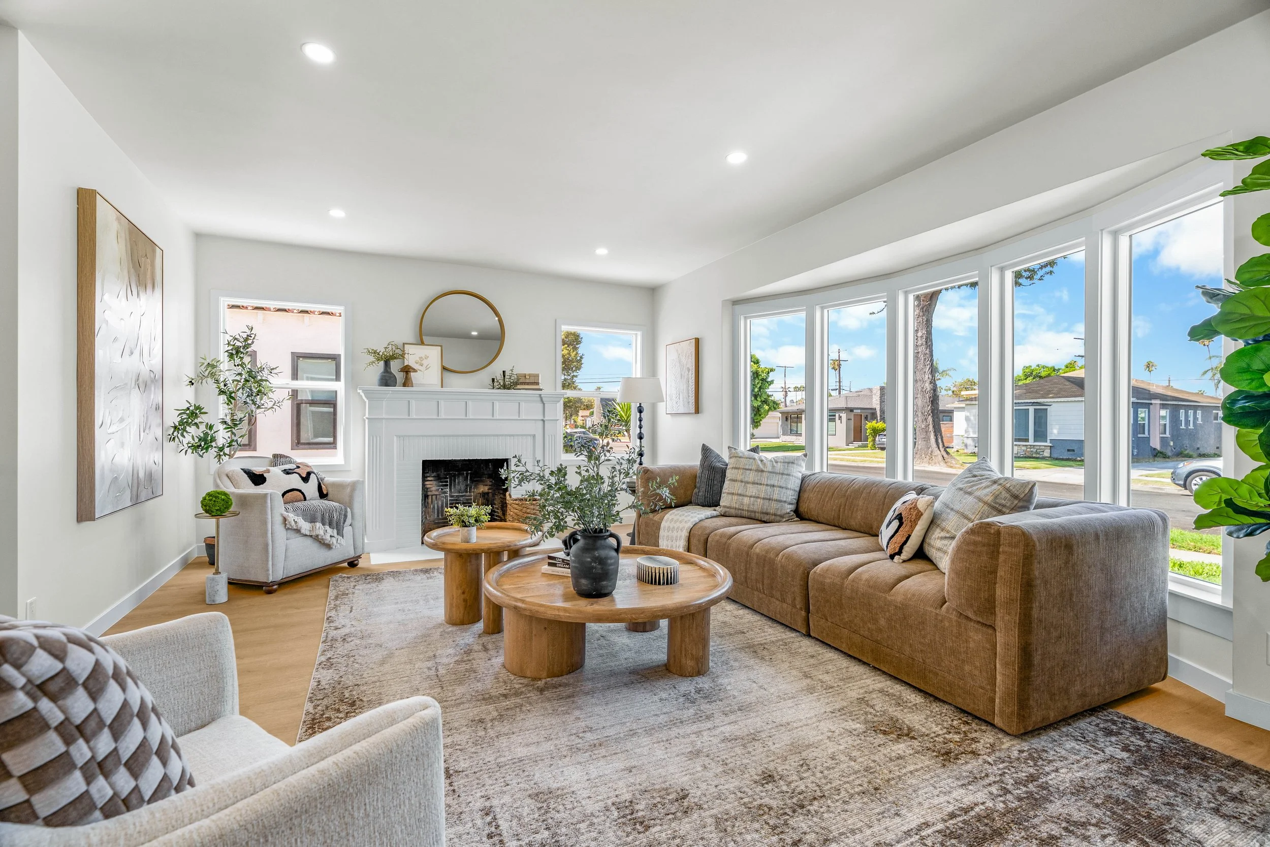 Bright living room with large windows, beige sofa, white armchair, wooden coffee tables, fireplace, plants, and modern decor.