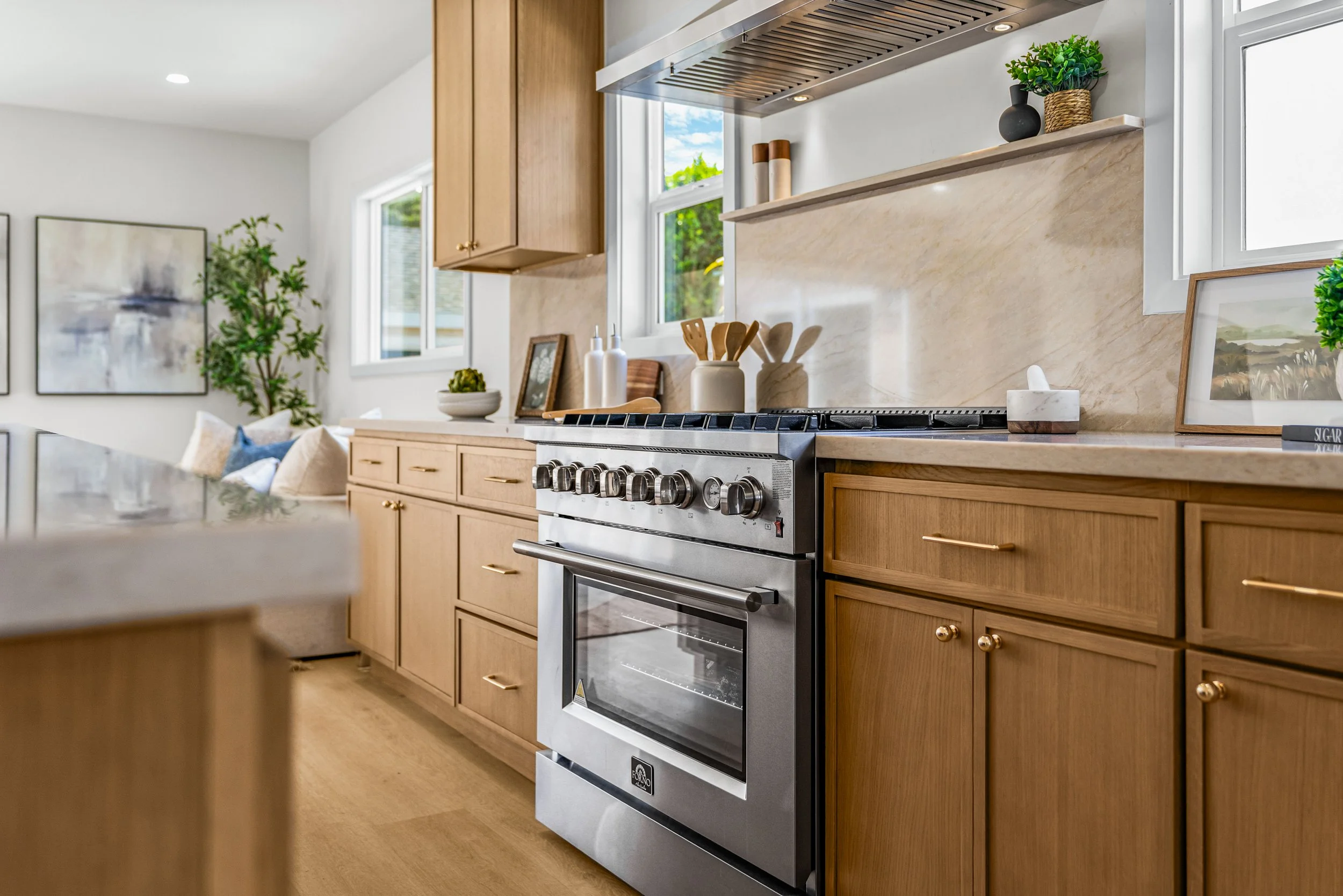 A modern kitchen with wooden cabinets, a stainless steel stove, and a beige marble backsplash. Decorative items including framed pictures and potted plants are on the countertops, with windows providing natural light.