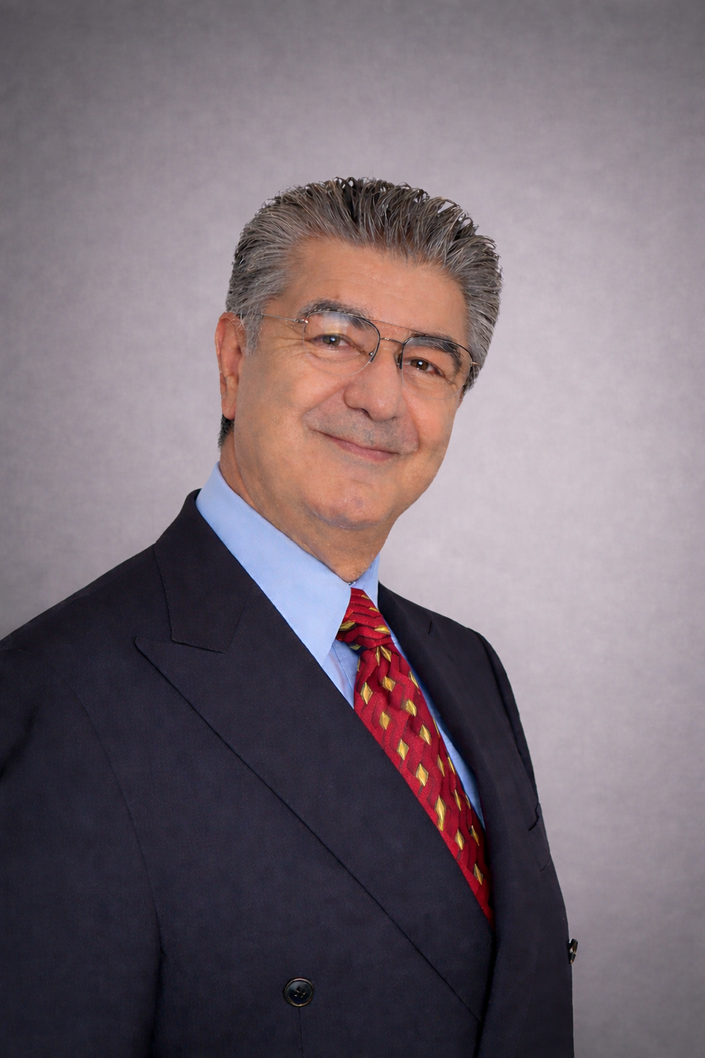 A middle-aged man with gray hair and glasses, wearing a dark suit, light blue shirt, and red tie, smiling against a plain gray background.
