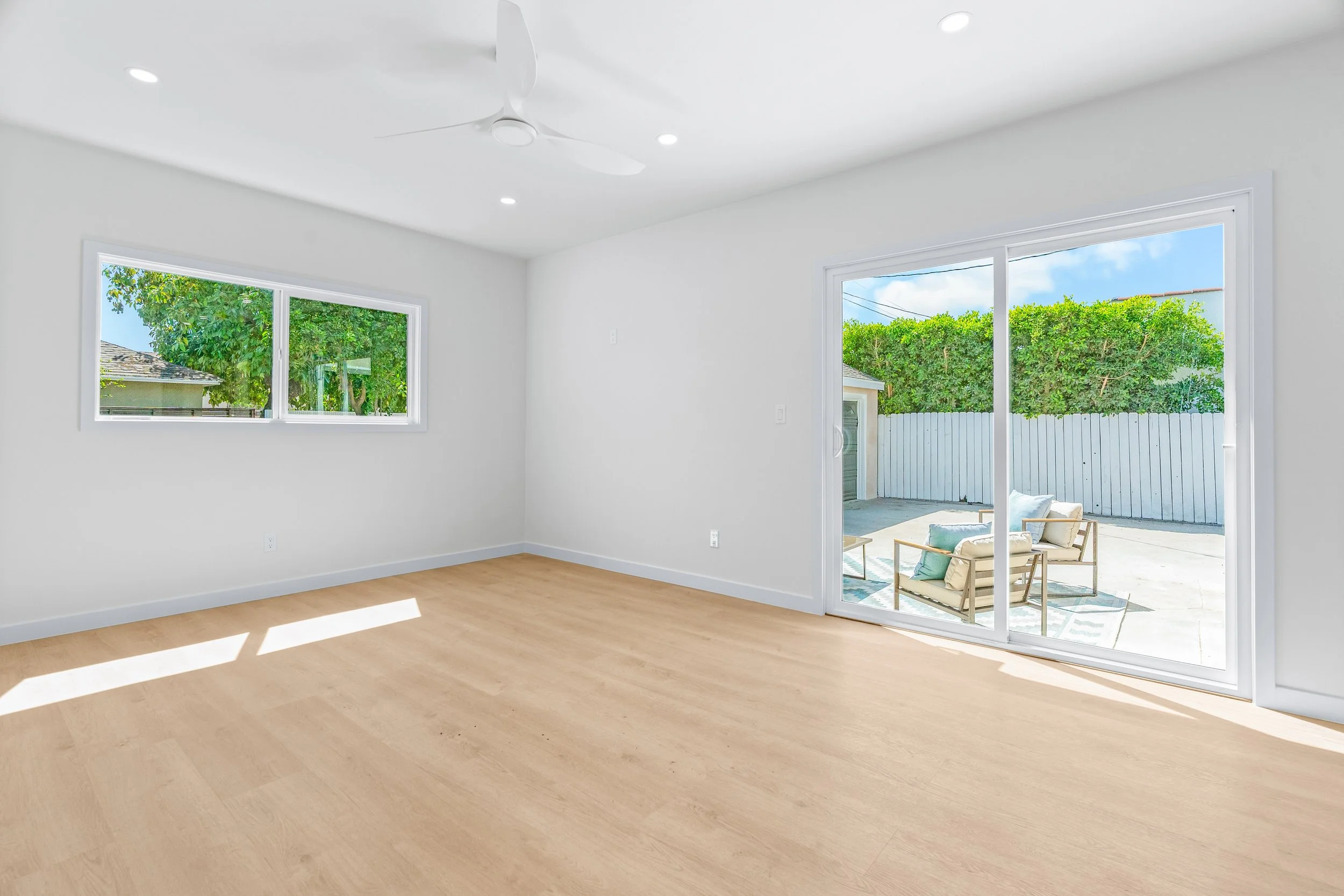 Empty living room with white walls, hardwood floor, large sliding glass door leading to patio, and two windows showing trees outside.