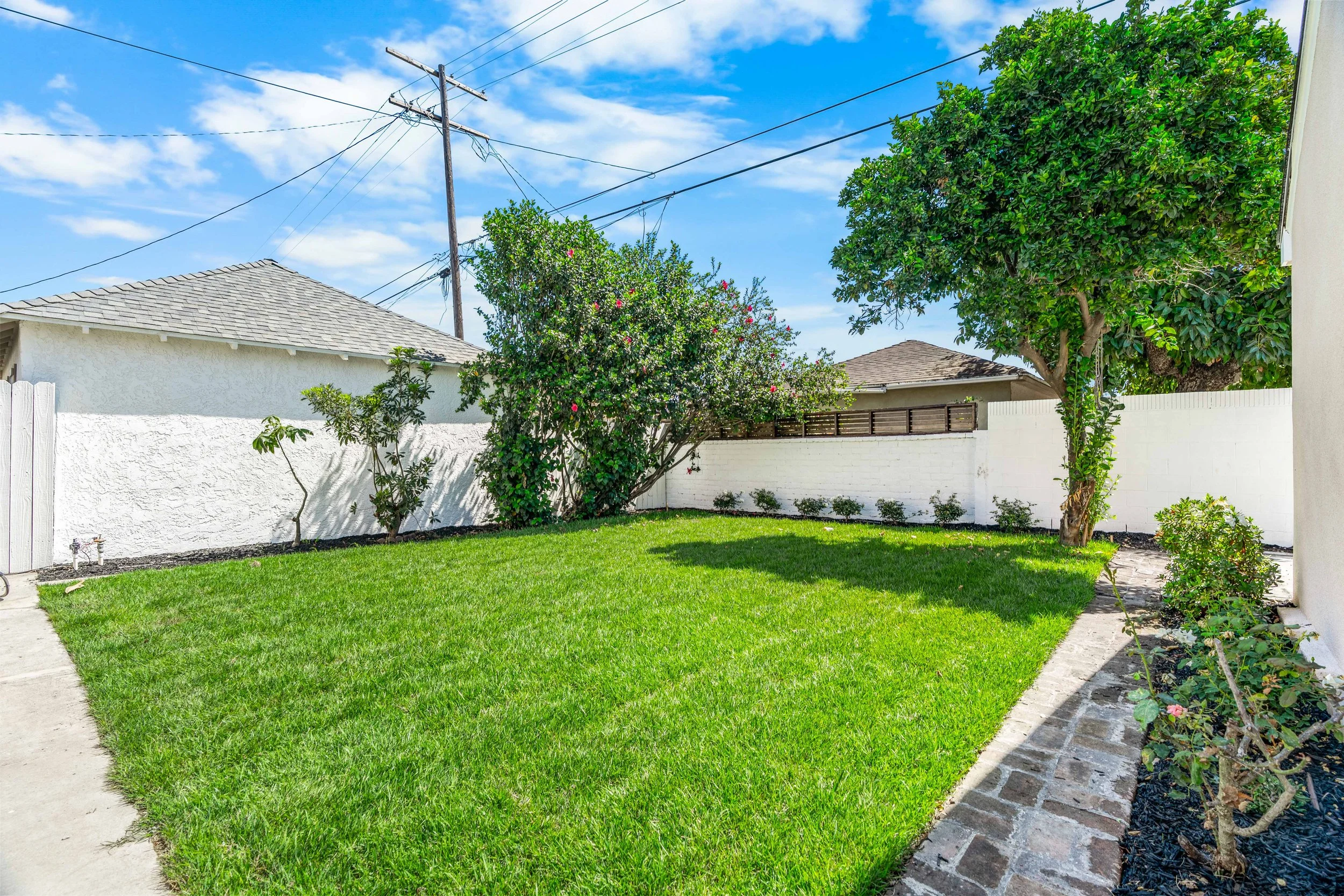 A backyard with a green lawn, trees, and shrubs, enclosed by a white brick wall. There is a concrete walkway along the right side and a garden bed with plants and flowers. The sky is blue with scattered clouds and an old wooden utility pole with power lines is visible.