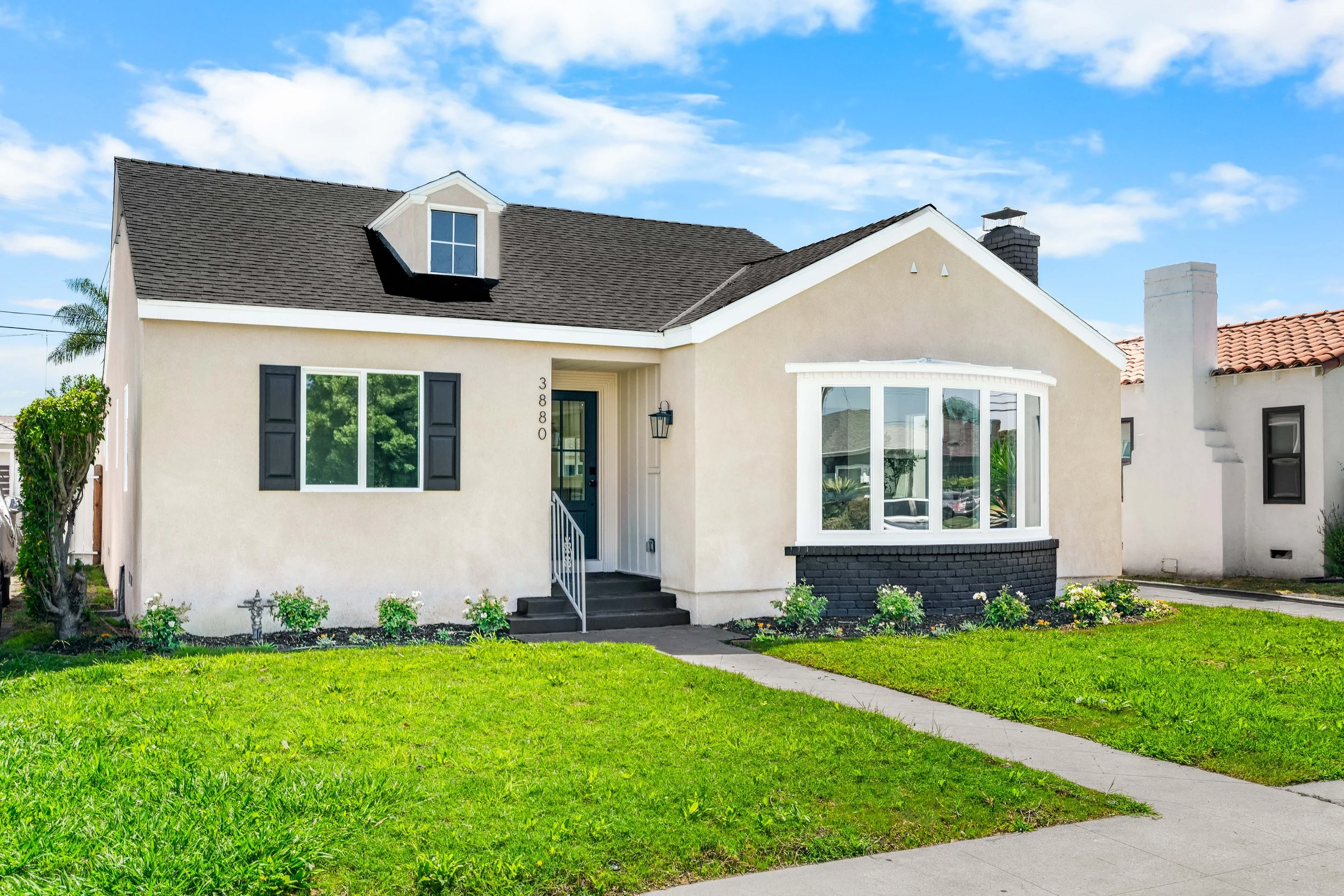 Front view of a beige house with black shutters, a bay window, and a small dormer window on the roof. There is a green lawn and a sidewalk in front of the house, with a partly cloudy blue sky in the background.