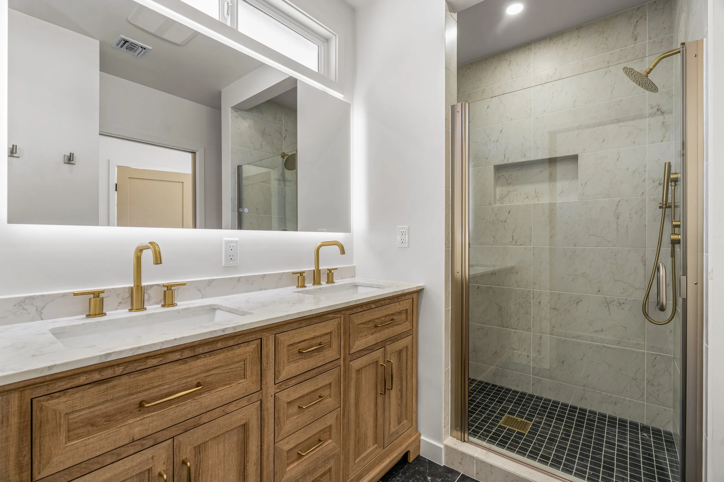 Modern bathroom with a wooden vanity with gold handle pulls, a white marble countertop with gold fixtures, a large illuminated mirror, a window, and a walk-in shower with beige marble tile walls, a glass door, a built-in niche, and a gold showerhead.