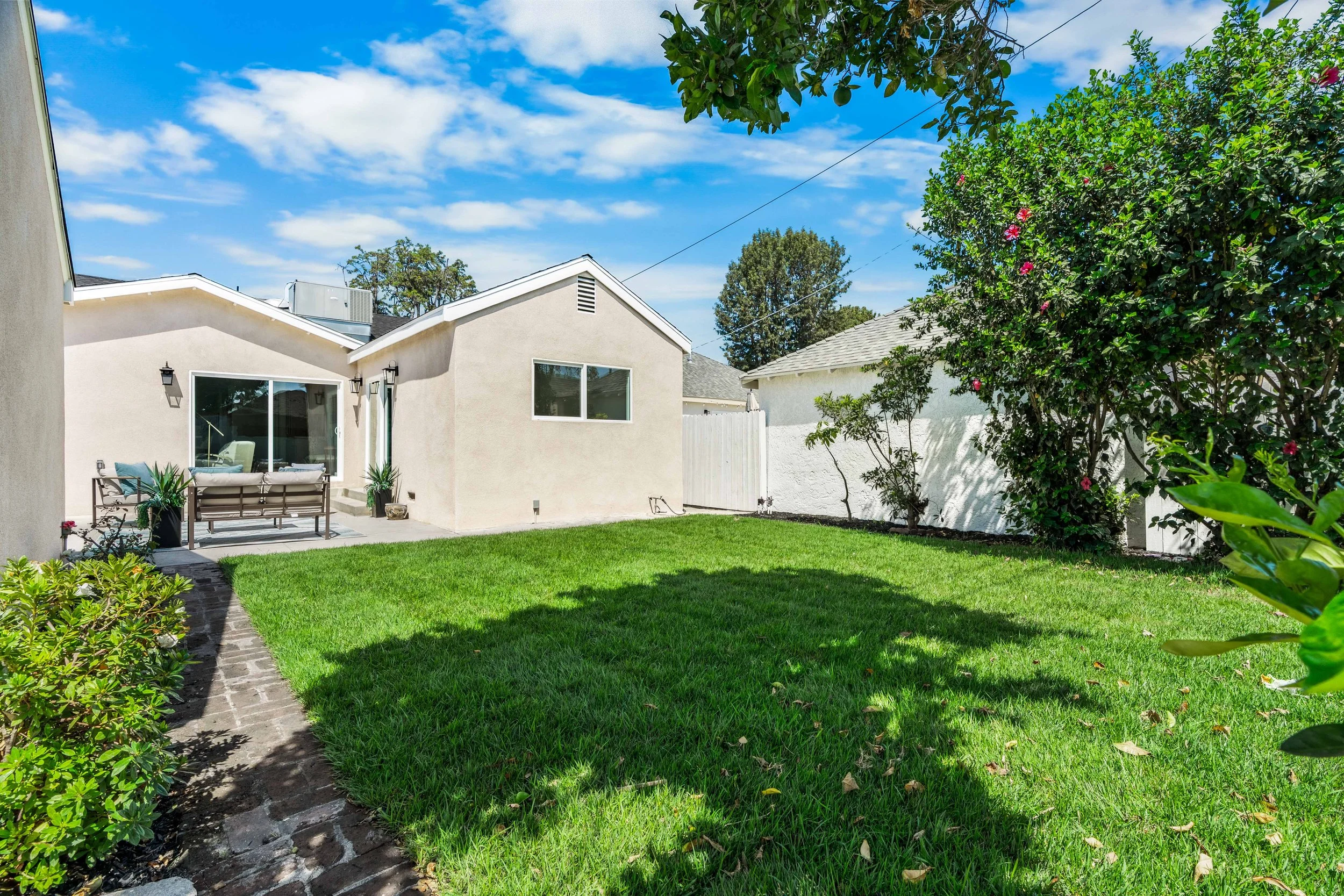 A backyard with a green lawn, a paved pathway along the left, a white stucco house with large windows and outdoor seating, trees, bushes with pink flowers, and a partly cloudy blue sky.