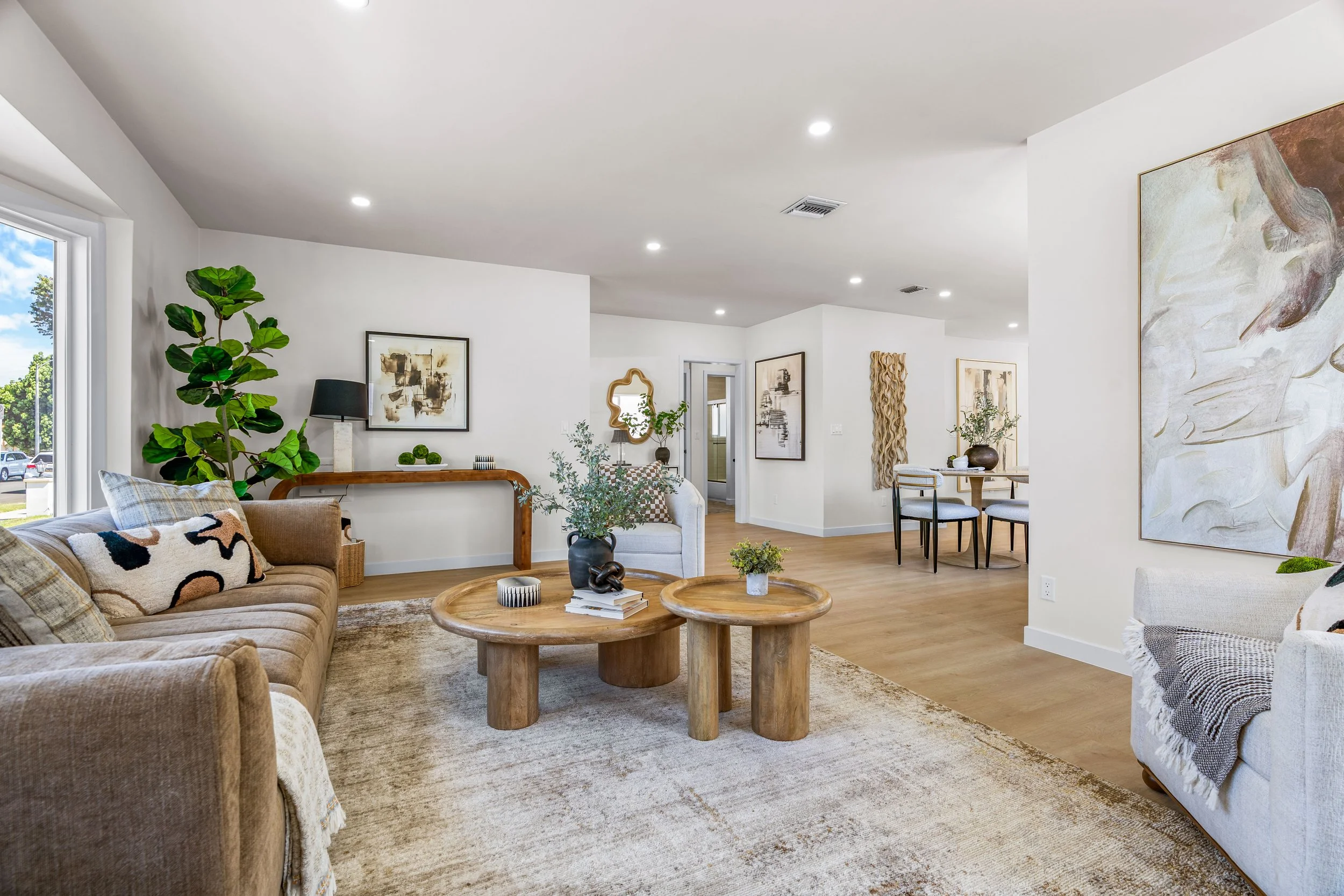A spacious and modern living room with beige sofas, a wooden coffee table, and decorative plants. Artwork decorates the white walls, and a large window lets in natural light. The room is open and inviting with a neutral color palette and contemporary decor.