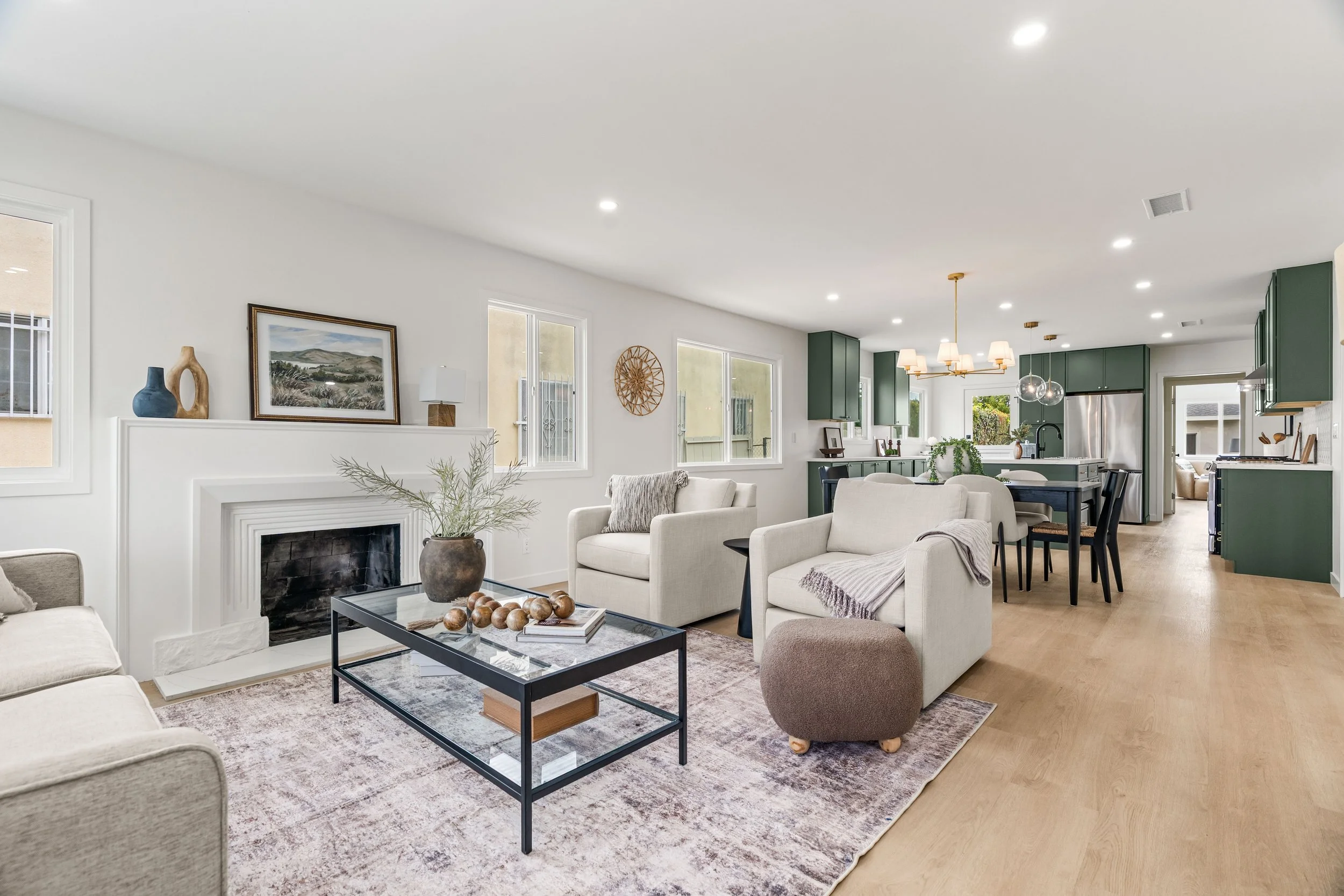 Open-concept living room and kitchen with white walls, wooden flooring, green kitchen cabinets, and modern lighting fixtures. Furniture includes white sofas, a black coffee table, and a patterned area rug.