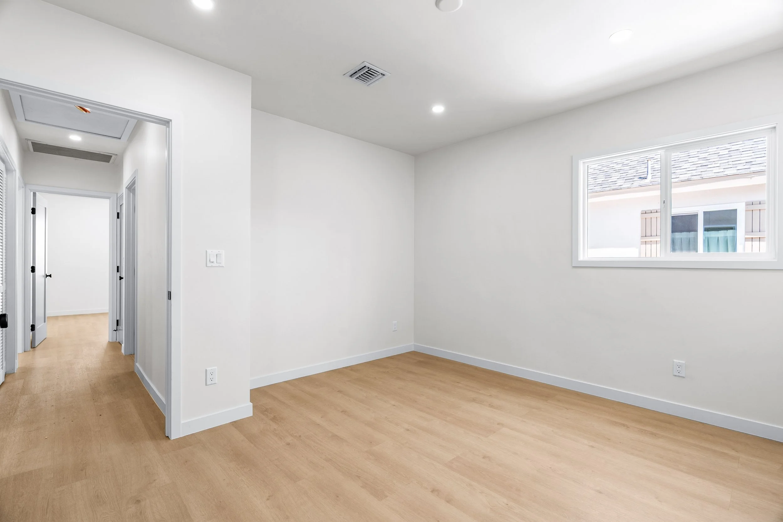 Empty room with white walls, wooden floor, a window, and doorways.