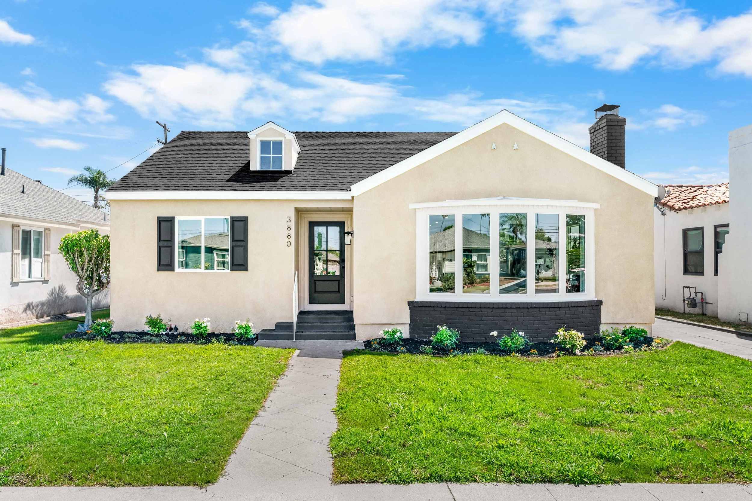 Front view of a cream-colored house with black shutters, a black front door, and a bay window with white trim, surrounded by a green lawn and bushes under a blue sky with scattered clouds.