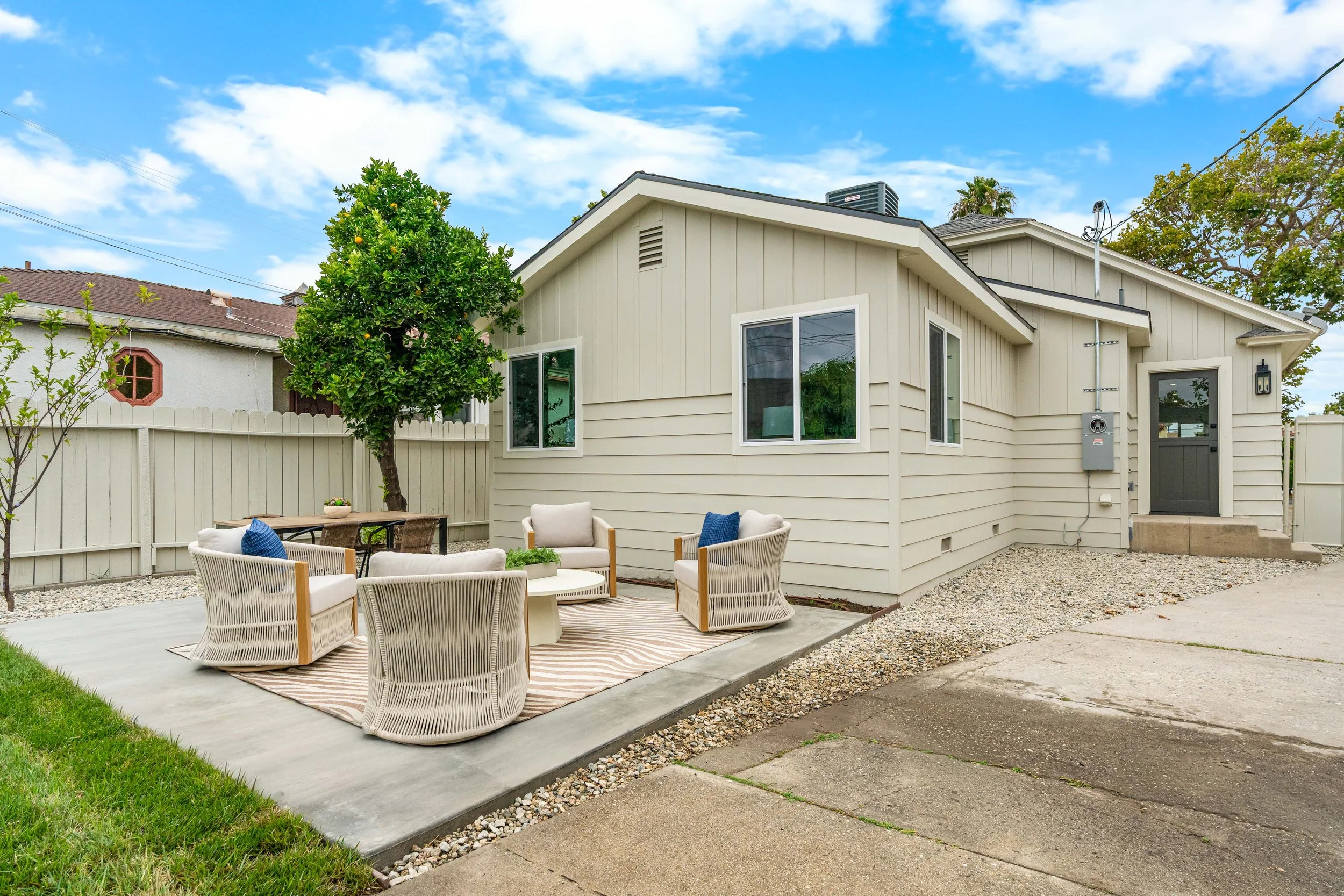 A backyard patio with outdoor furniture including a striped rug, cushioned chairs, and a small table, enclosed by a white wooden fence, with a house featuring beige siding, multiple windows, and a tree with green leaves under a partly cloudy sky.