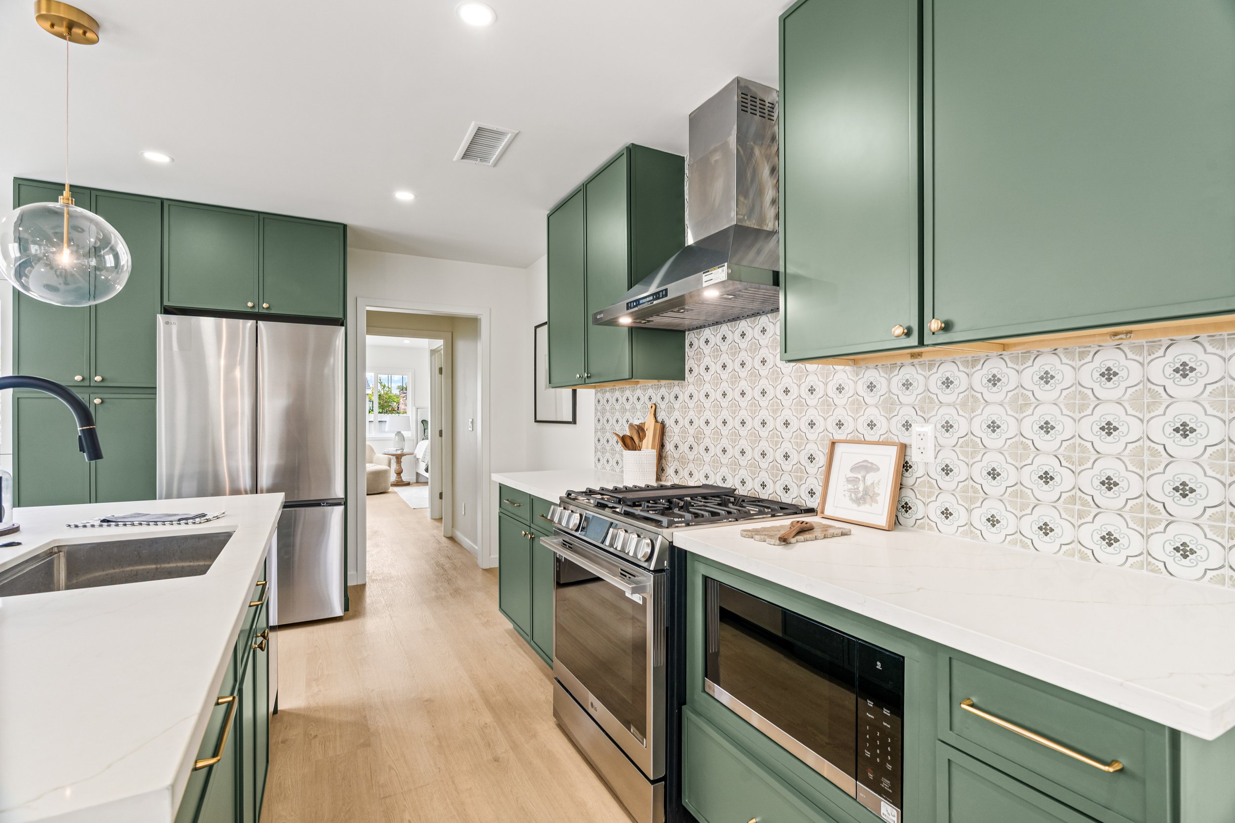 Modern kitchen with green cabinets, white countertops, stainless steel appliances, decorative tiled backsplash, and a view into another room with natural light.