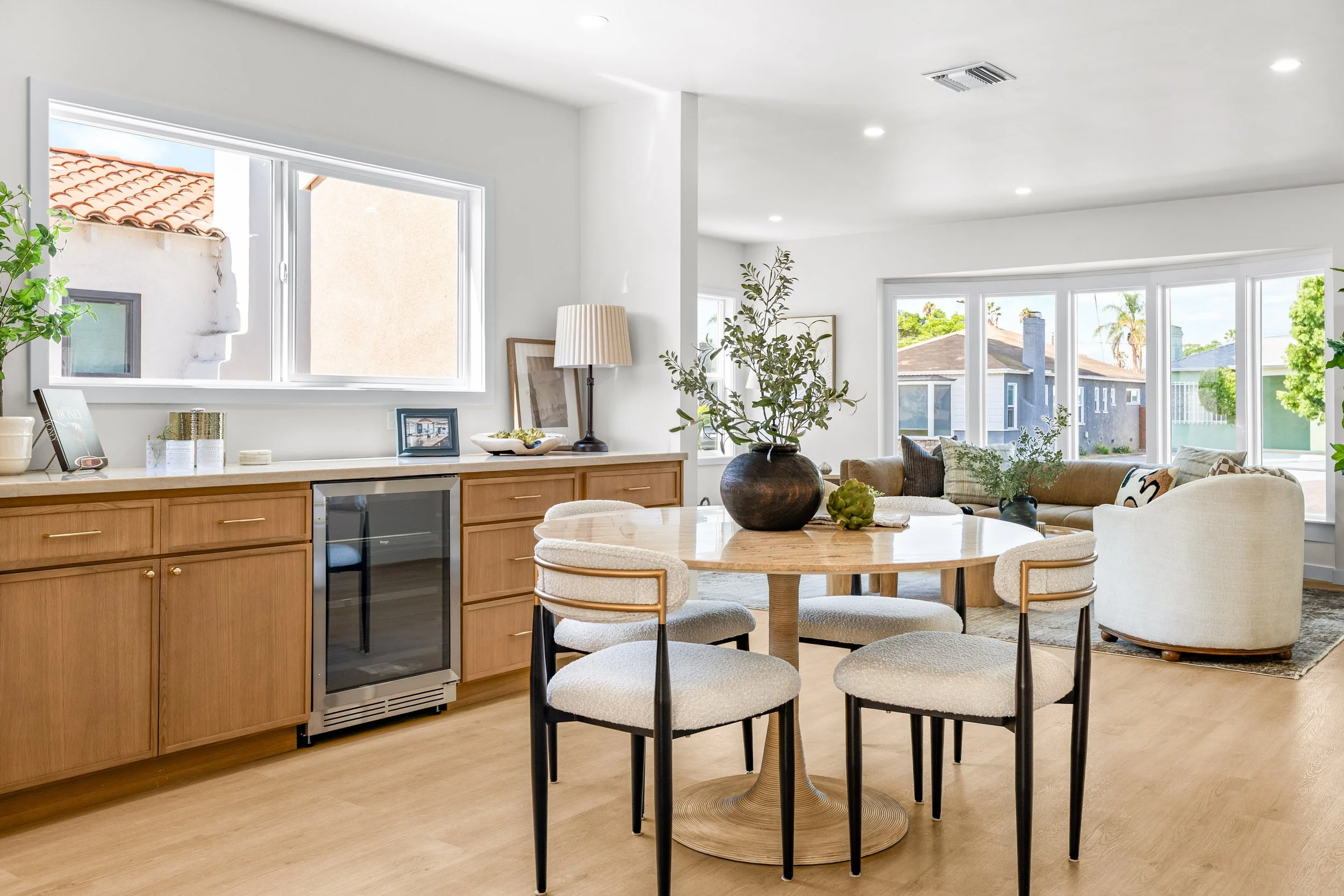 Bright open living room with large windows, beige couches, a round wooden dining table with four upholstered chairs, houseplants, and a breakfast bar with wooden cabinets and small decor items.