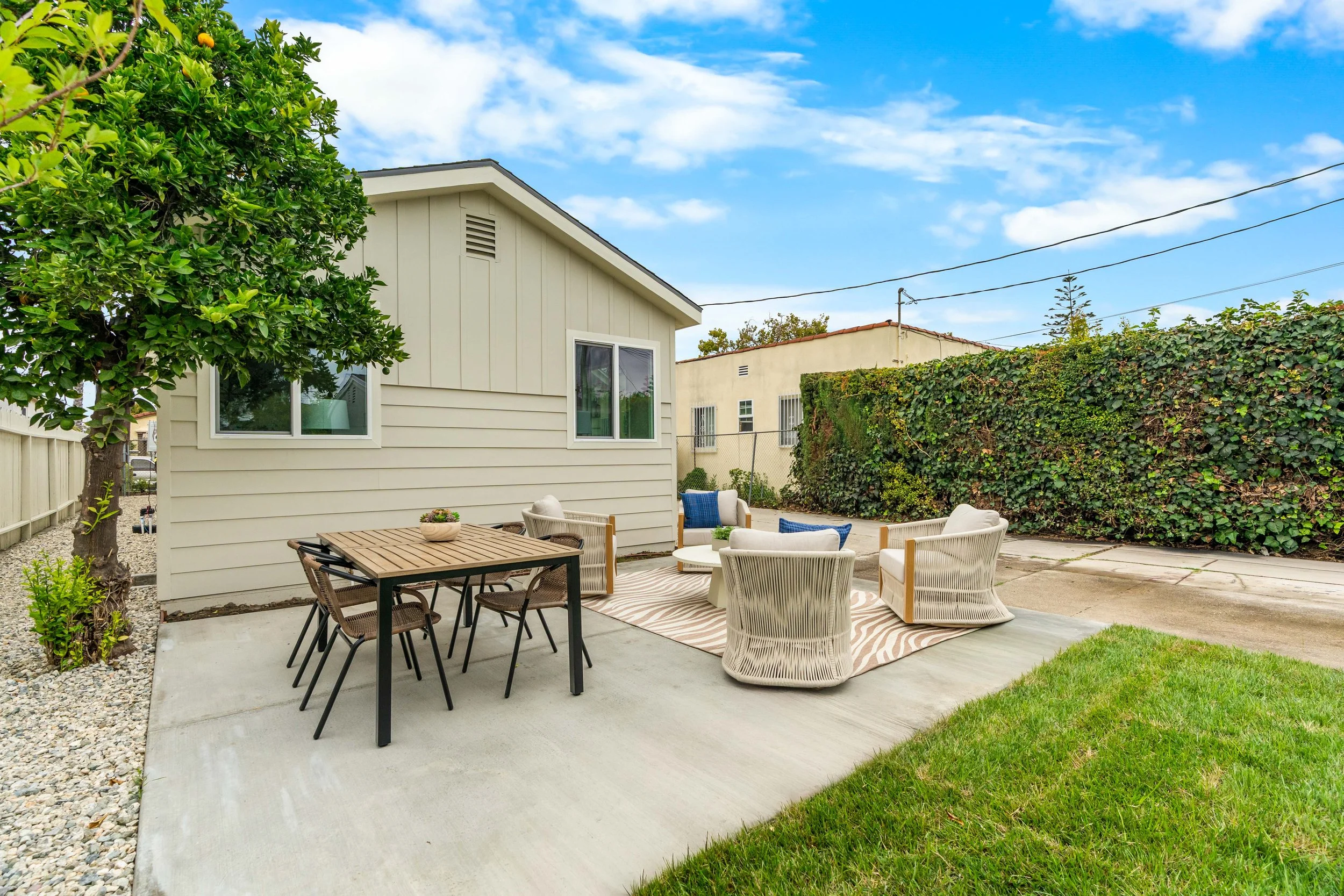 A backyard patio area with outdoor furniture, including a table and multiple chairs, next to a light-colored house with large windows. There is a tree with green foliage on the left and a high hedge on the right, under a partly cloudy blue sky.
