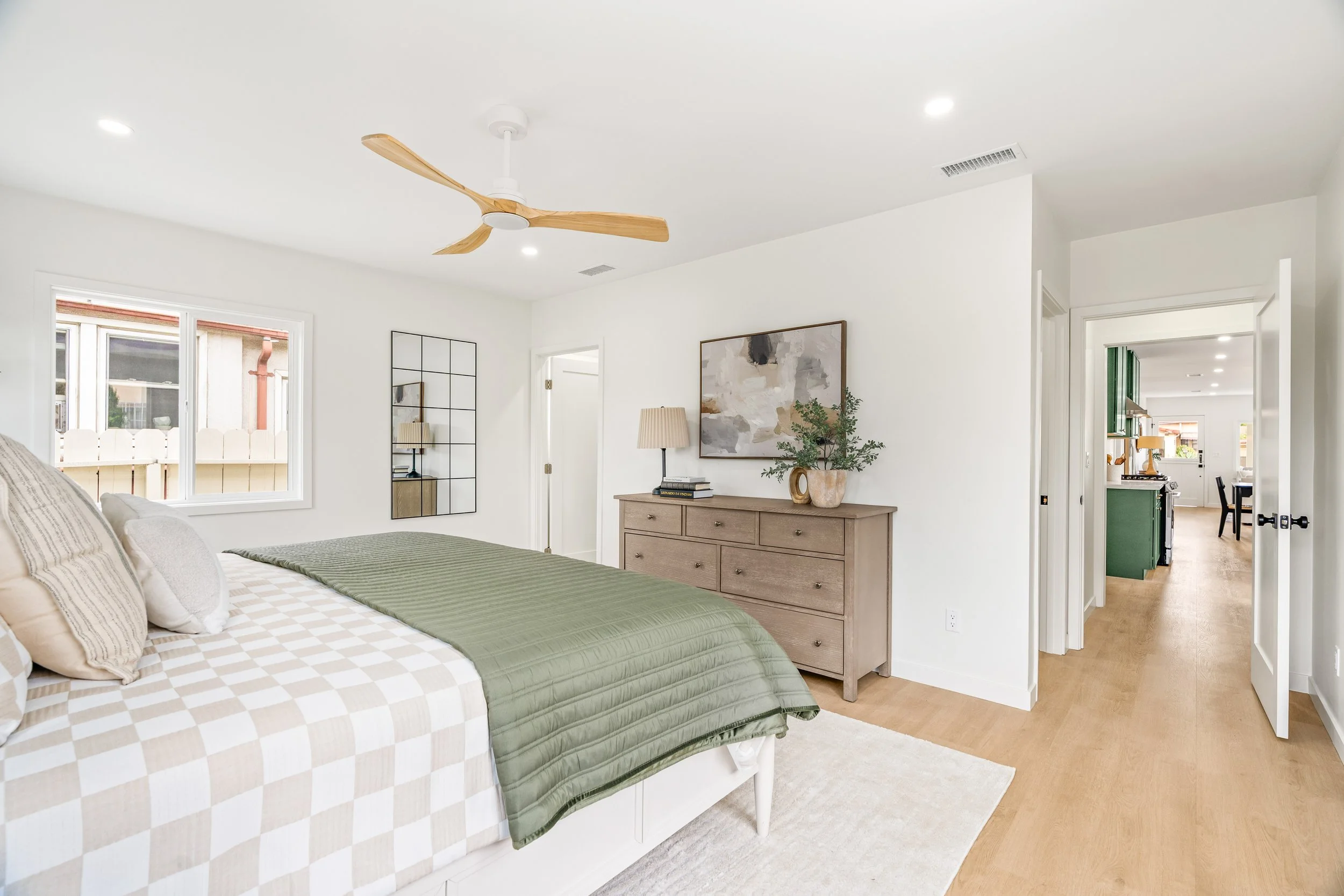 A bedroom with a bed featuring a checkered beige and white bedspread, multiple pillows, a wooden dresser with a plant and picture frame on it, a mirror, and a window with white blinds. A hallway leads to a kitchen area with green cabinetry.
