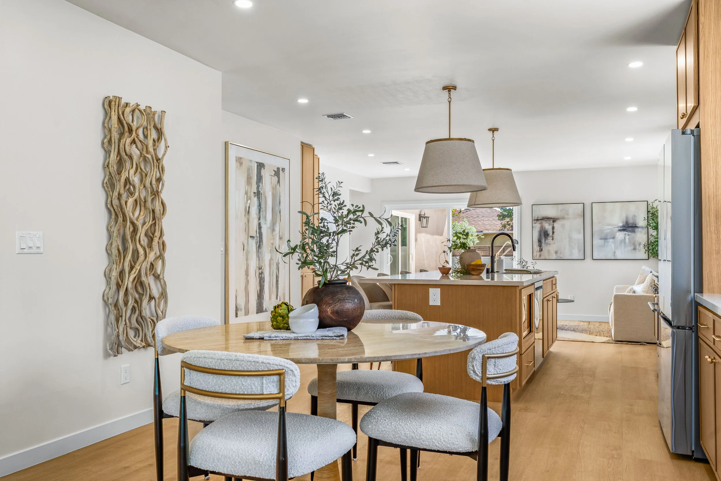 Interior of a modern home with a dining area and kitchen. The dining table with white upholstered chairs is in the foreground, decorated with a large ceramic vase, a small white bowl, and a pineapple. The kitchen in the background has wooden cabinetry, pendant lights, and a window showing an outdoor view. Wall art and a textured wall hanging are visible on the white walls. Hardwood floors and recessed ceiling lights complete the space.
