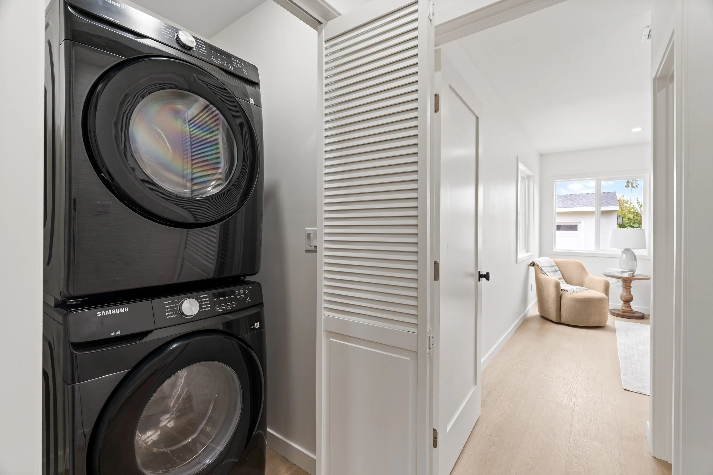 A laundry closet with a stacked black Samsung washer and dryer behind a white louvered door. Adjacent to the laundry space is a bright living area with a window, a beige armchair, a small wooden side table, and a white lamp.
