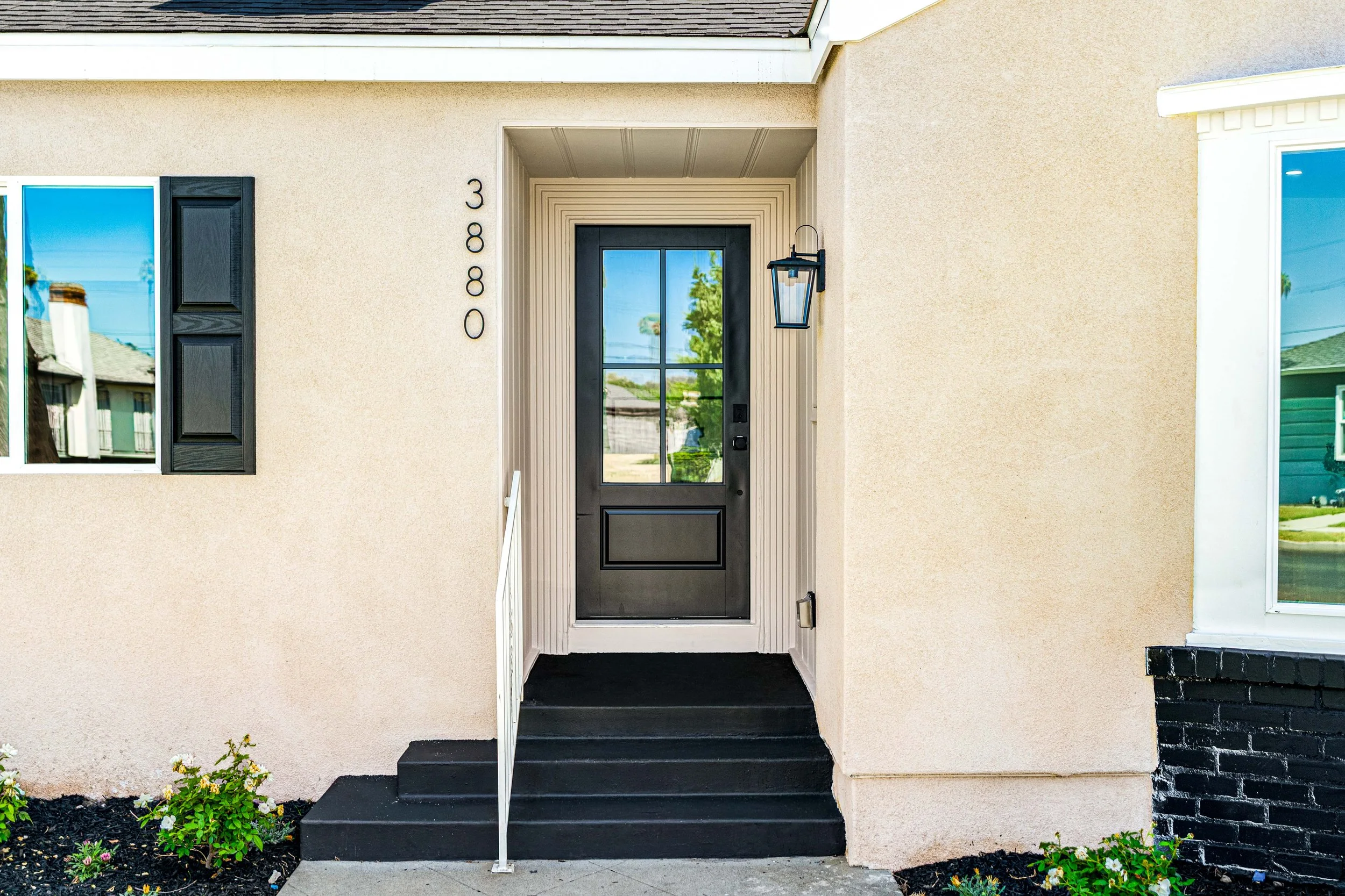 Front view of a house entrance with a black door, black shuttered window, and a lantern-style light fixture, with flower beds on each side.