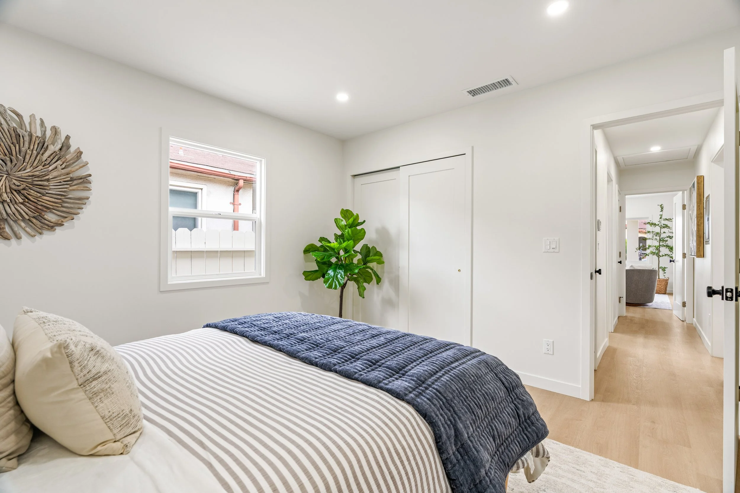 A bedroom with white walls and a bed with striped bedding, pillows, and a dark blue quilt. There is a green potted plant near the window, a decorative wall piece resembling a sun or flower made of driftwood, and a doorway leading to another room with