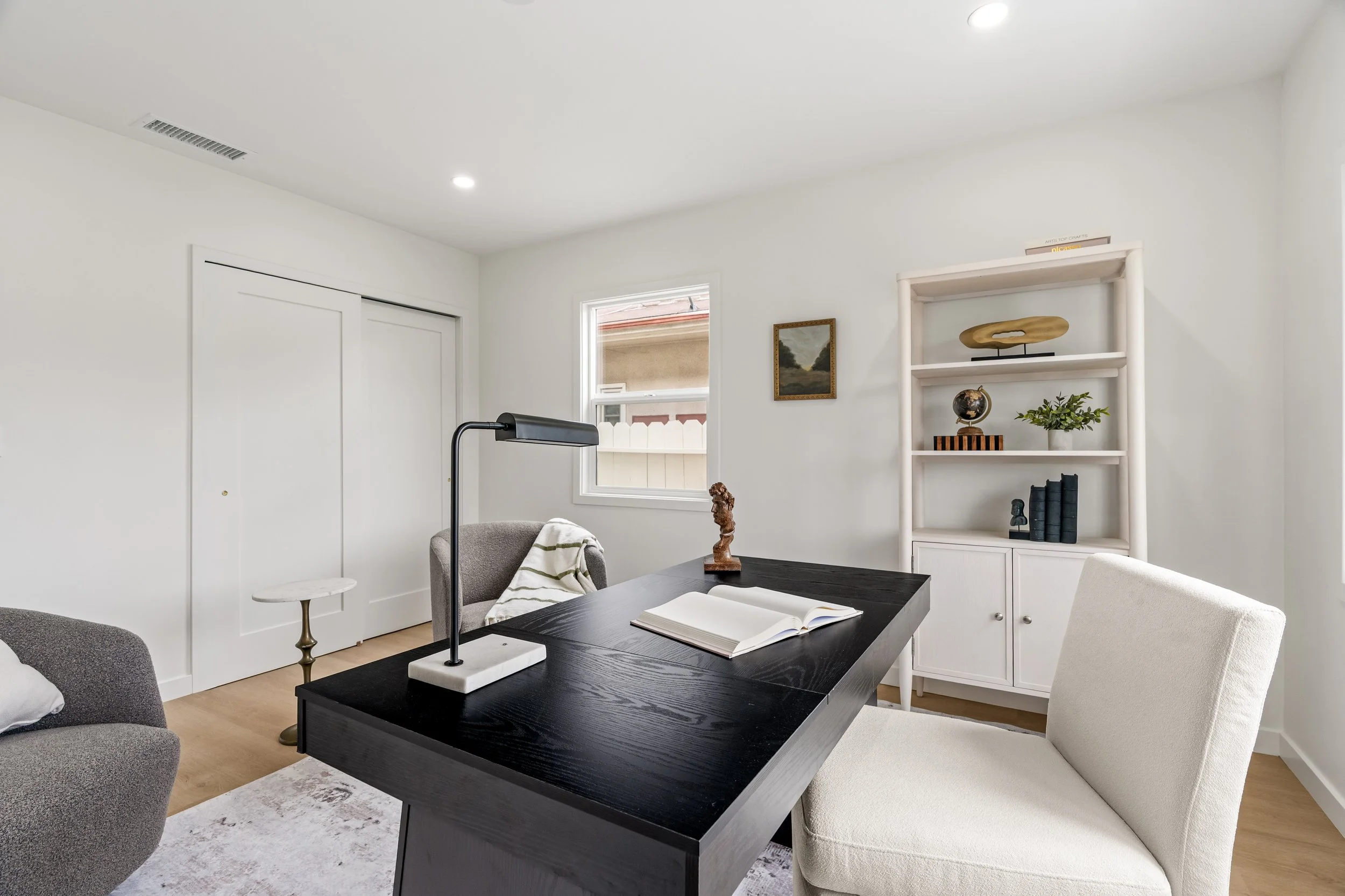 A modern, minimalist office or study room with white walls and a black desk. The room contains two chairs, a gray and a white, a bookshelf decorated with a wooden sculpture, a globe, books, and a plant. There is a small window and recessed ceiling li