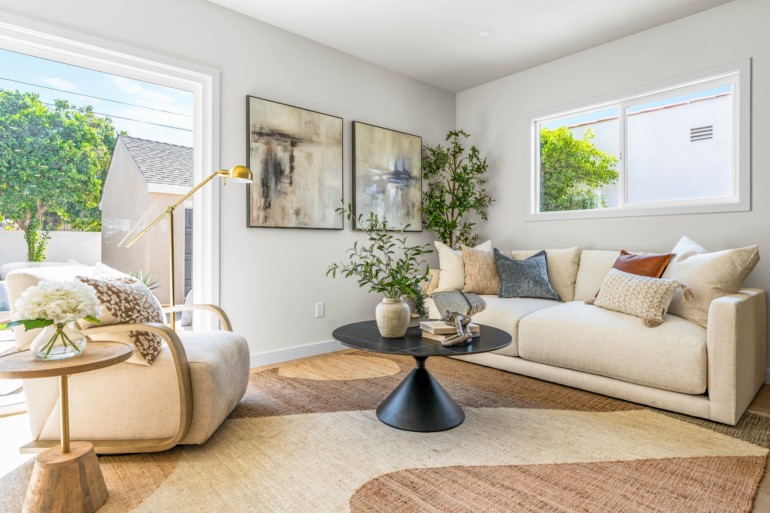 Living room with large beige sectional sofa with pillows, black round coffee table with books, ceramic vase, and decorative objects, wall art, tall potted plant, and windows showing outdoor greenery, natural light, and minimalist decor