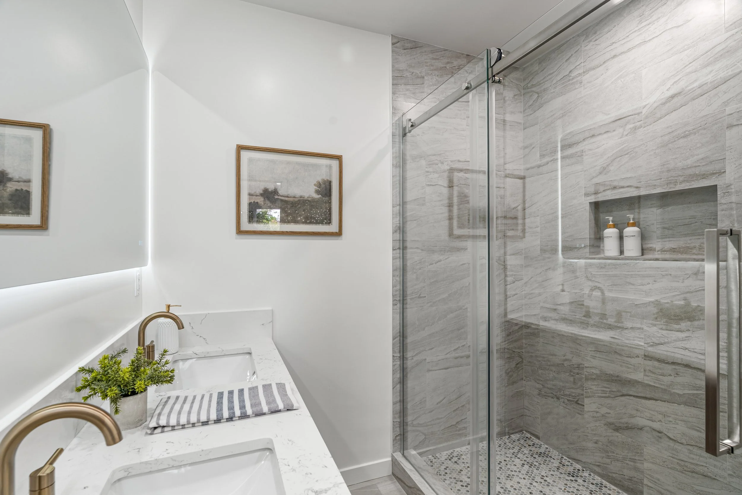 A modern bathroom with a white vanity, sink, and gold faucet, decorated with a small plant and striped towel, next to a glass-enclosed shower with marble walls and built-in shelves with shampoo and conditioner bottles.