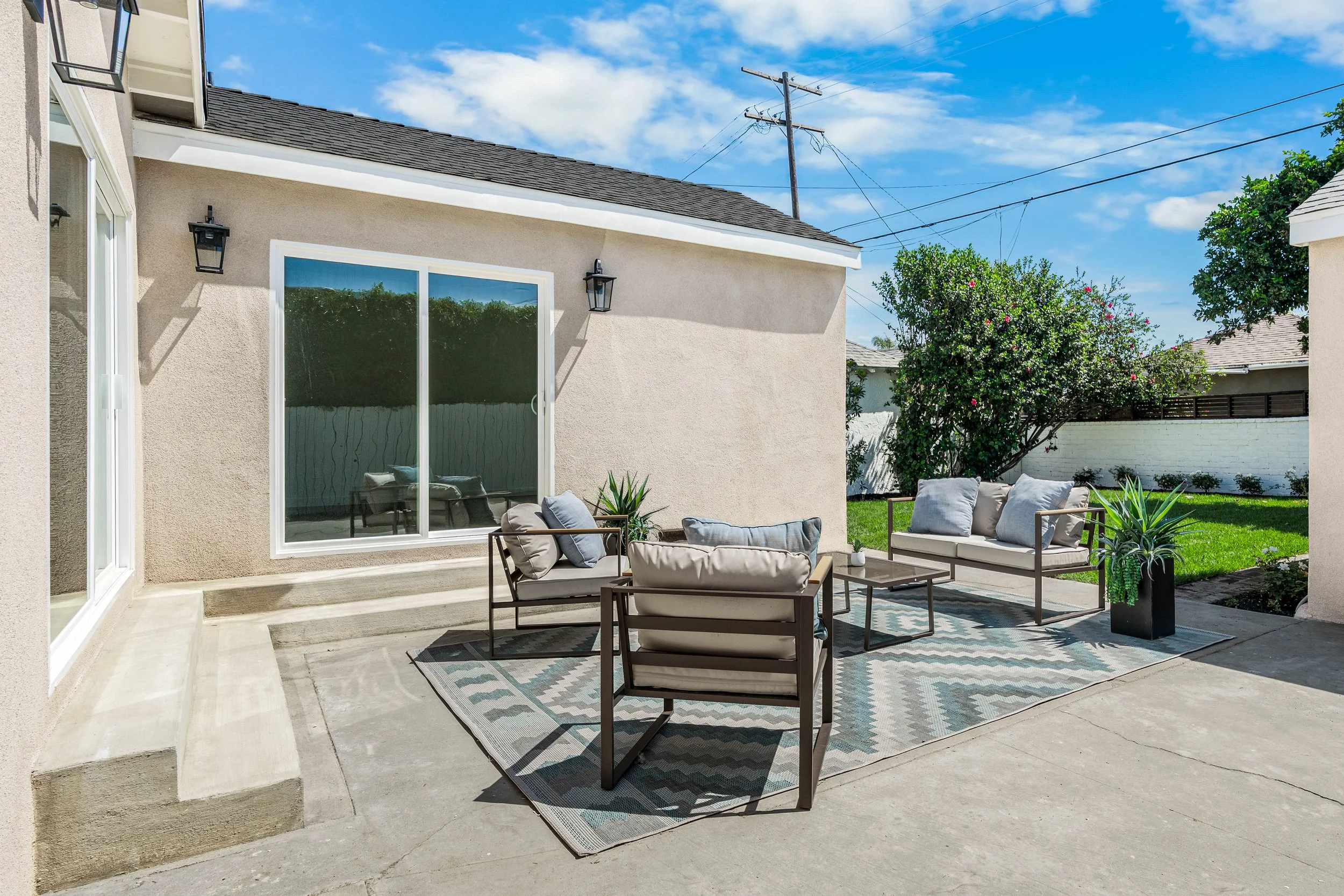 Backyard patio with outdoor furniture including chairs and a coffee table, area rug, potted plants, and a view of a house wall with large windows, outdoor lighting, and a fenced yard with trees under blue sky.