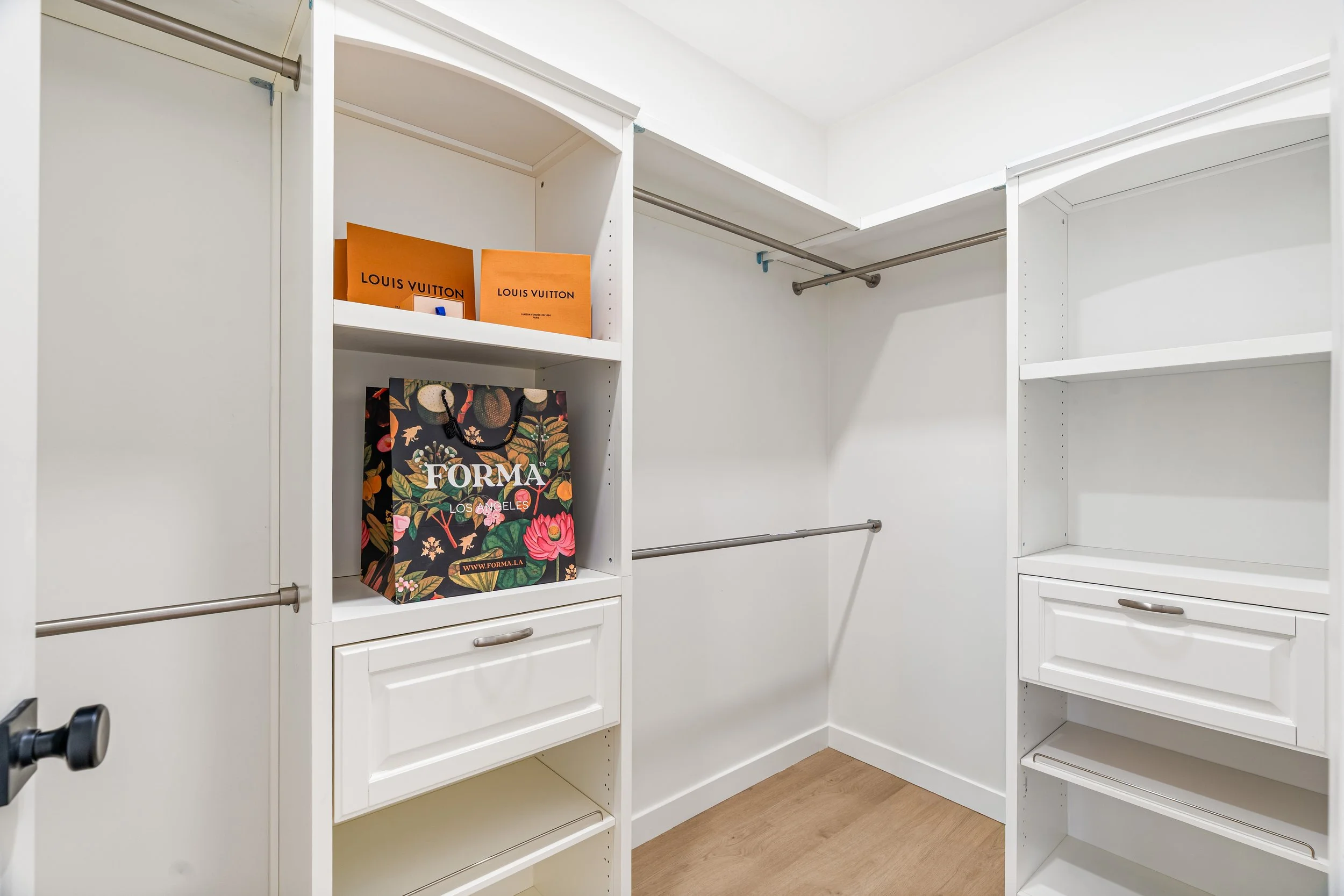 A walk-in closet with white shelves, some with hanging rods, and a few decor items including an orange Louis Vuitton shopping bag and a colorful shopping bag with floral designs.