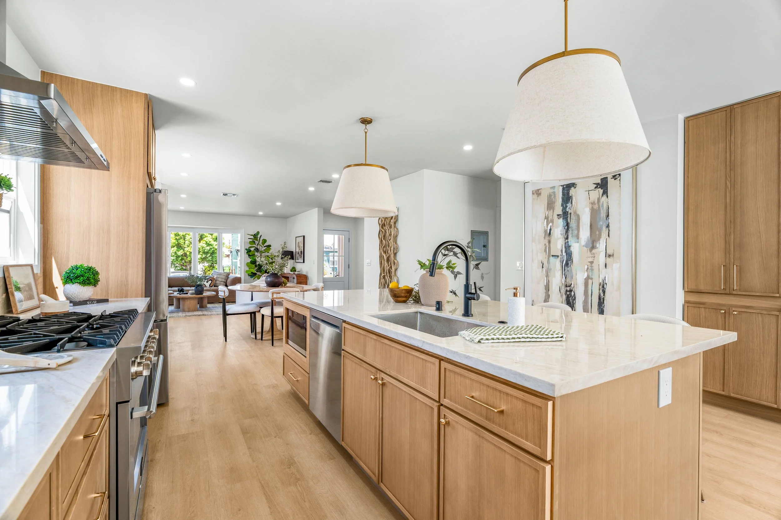 Modern open-concept kitchen with light wood cabinets, white marble countertops, and large pendant lights, overlooking a living room with large windows and greenery outside.