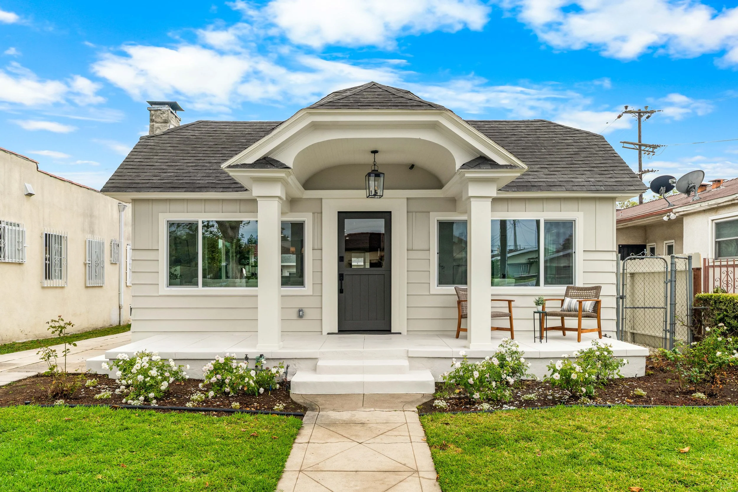 Front view of a small, white house with a dark door, large windows, and a front porch with a lantern hanging from the ceiling. There are two chairs and a small table on the porch, with a garden bed filled with white flowers in front. The house has a 