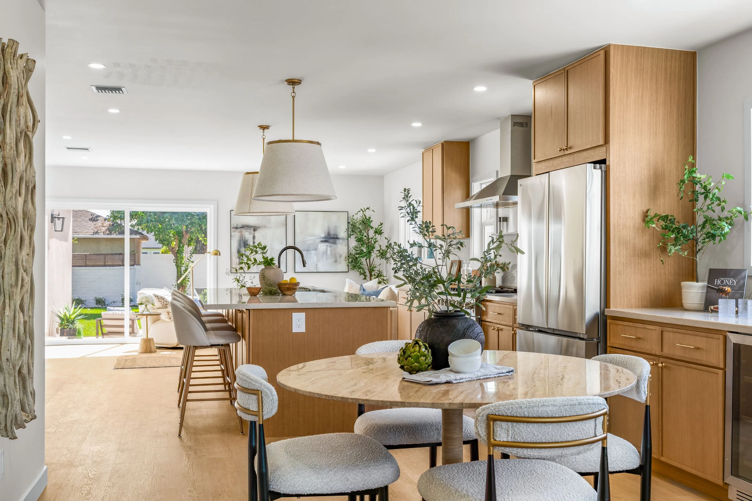 Open concept kitchen and living room with wooden cabinets, stainless steel refrigerator, white walls, pendant lighting, round dining table with chairs, plants, and large window with view of backyard.